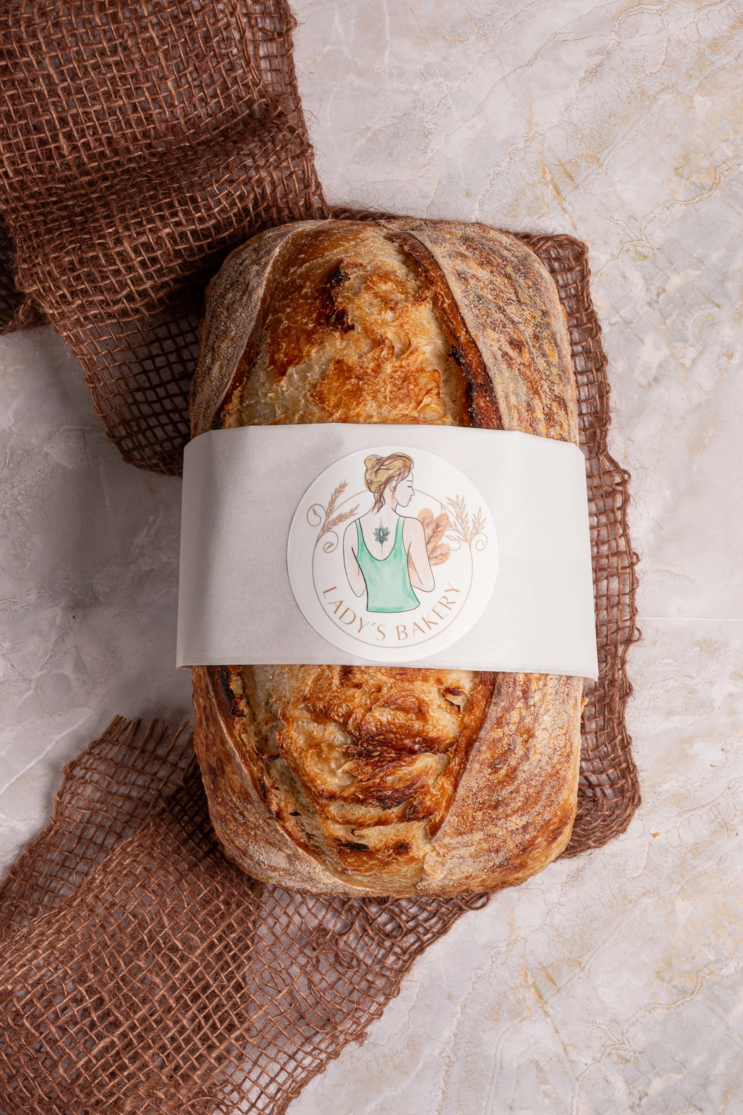 Loaf of bread wrapped in bakery paper with label showing a woman in a green apron, on a brown cloth, on a light-colored marble surface.