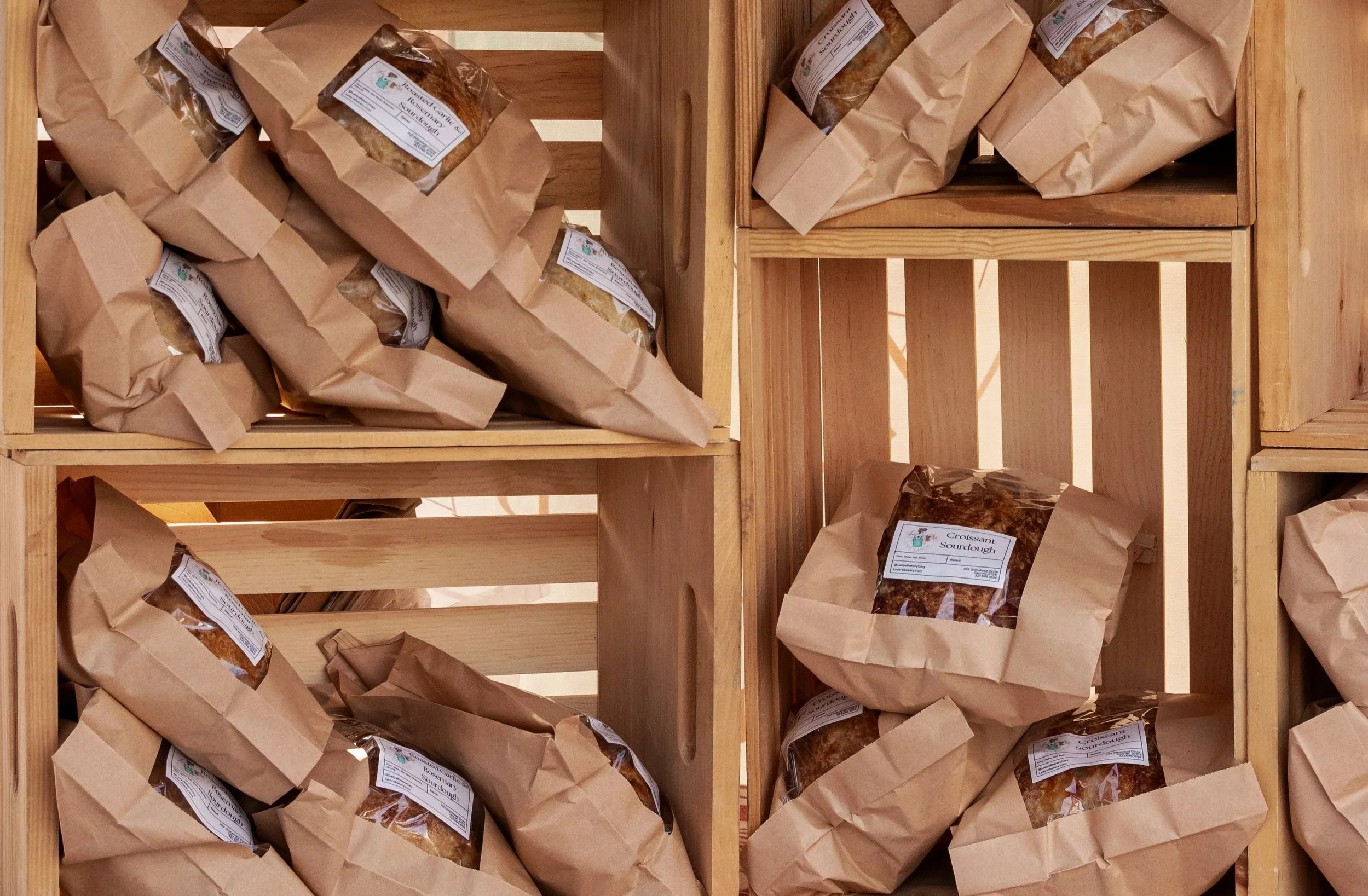 Wooden shelves filled with brown sourdough loaves in brown paper storage bags labeled 'Croissant Sourdough.'