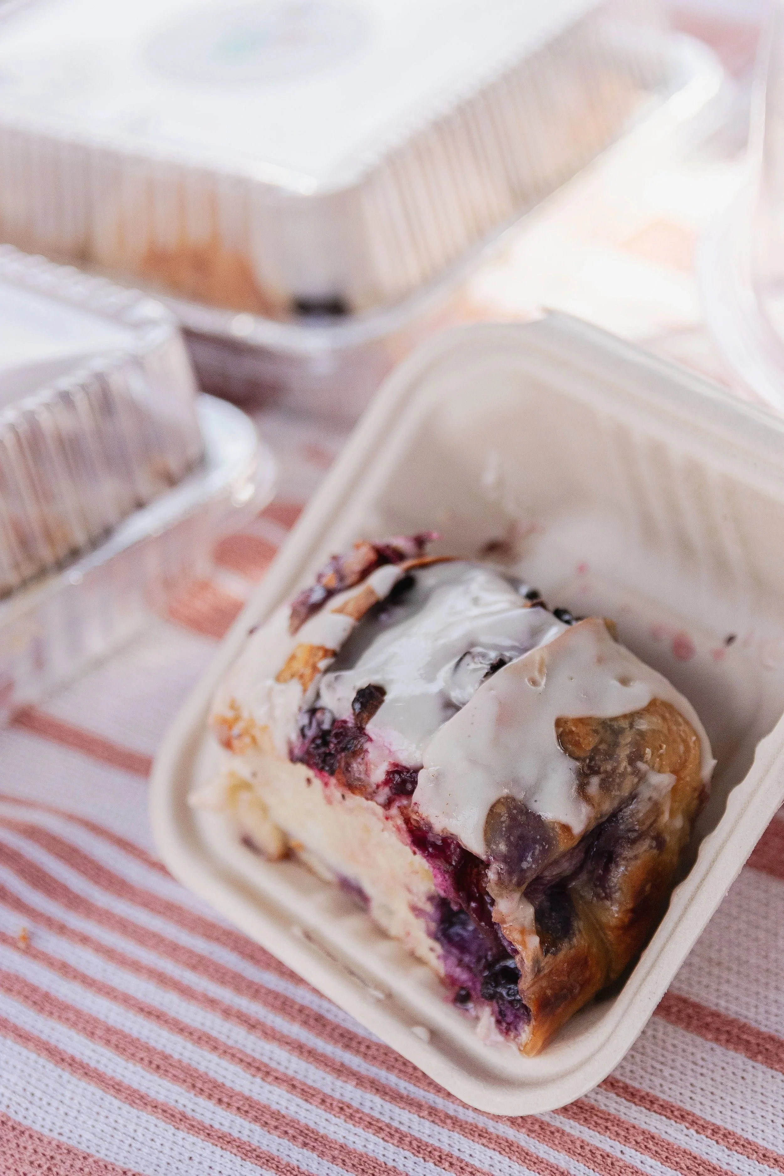 A piece of blueberry cinnamon roll with white icing on top in a disposable container, with several other containers around it.