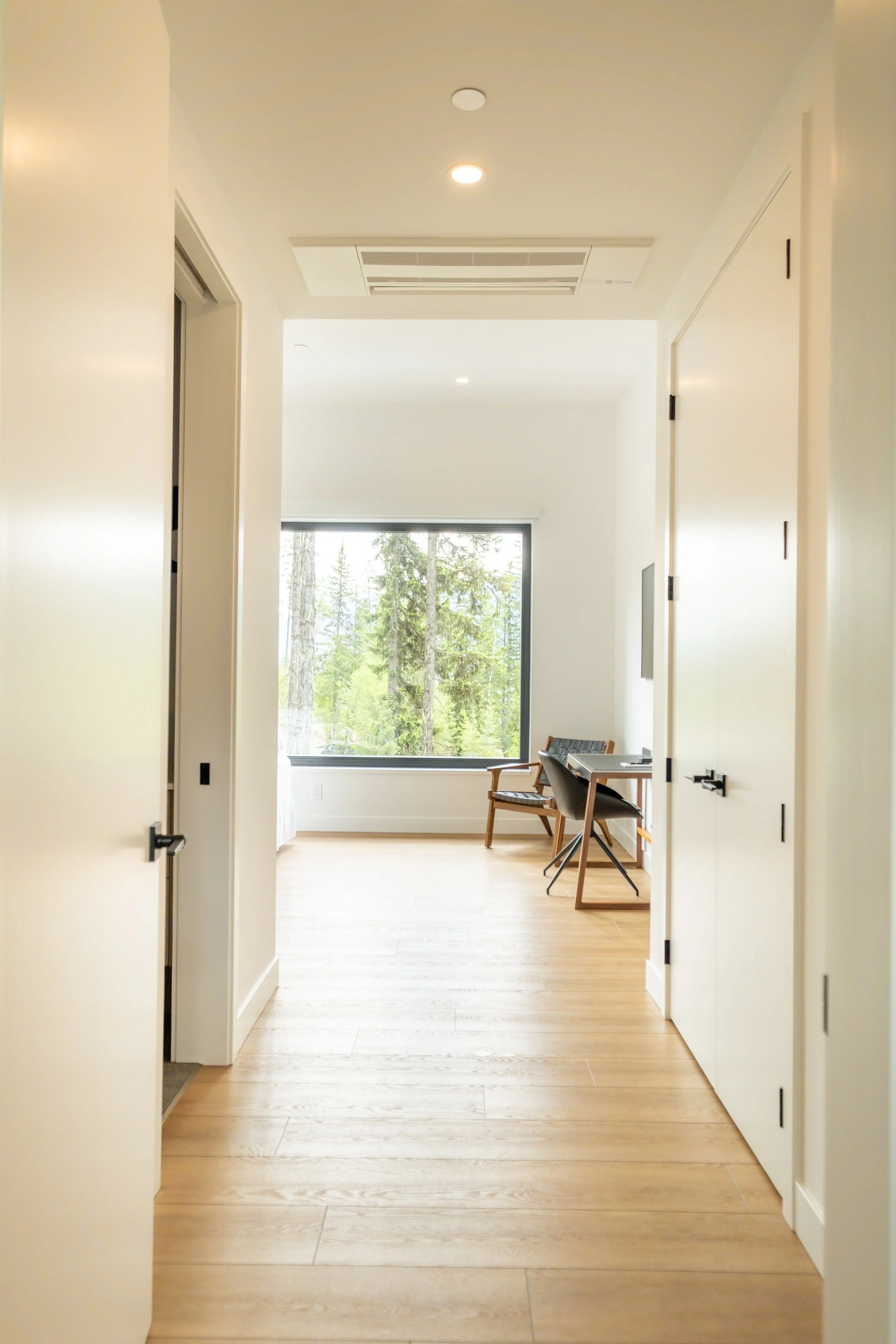 A bright hallway with light wood flooring, white walls and ceiling, leading to a room with large window showing trees outside. Two chairs and a small desk are visible in the room.
