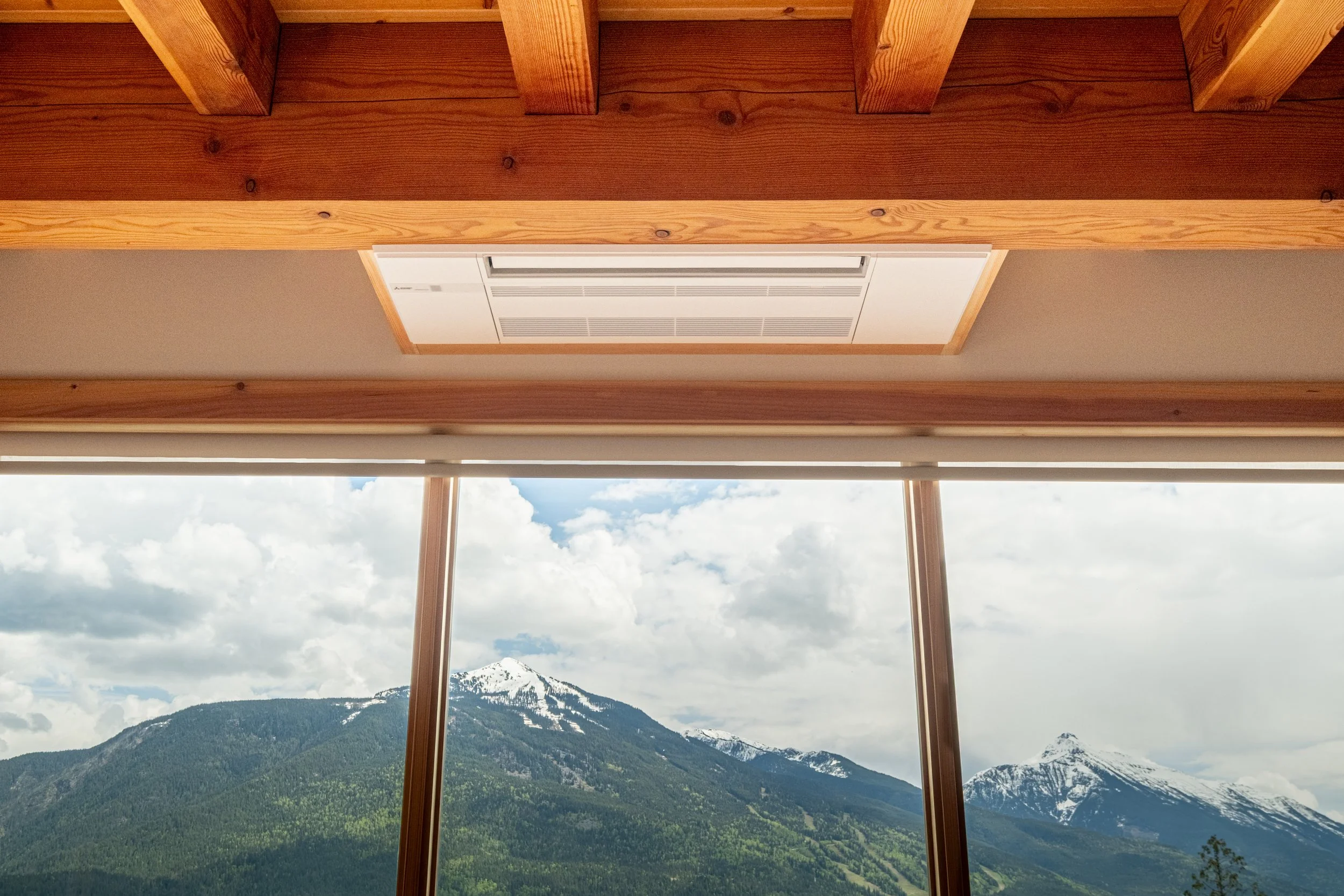 Indoor view showing a large window with a mountain landscape outside, including snow-capped peaks and green slopes, with parts of a wooden ceiling and a ceiling air conditioning unit visible.
