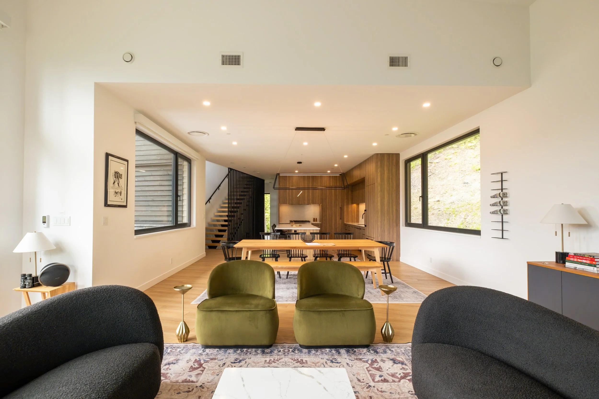 A spacious, modern living room with black and olive green armchairs, a wooden dining table with black chairs, a kitchen area with wooden cabinetry, large windows, and a staircase in the background.