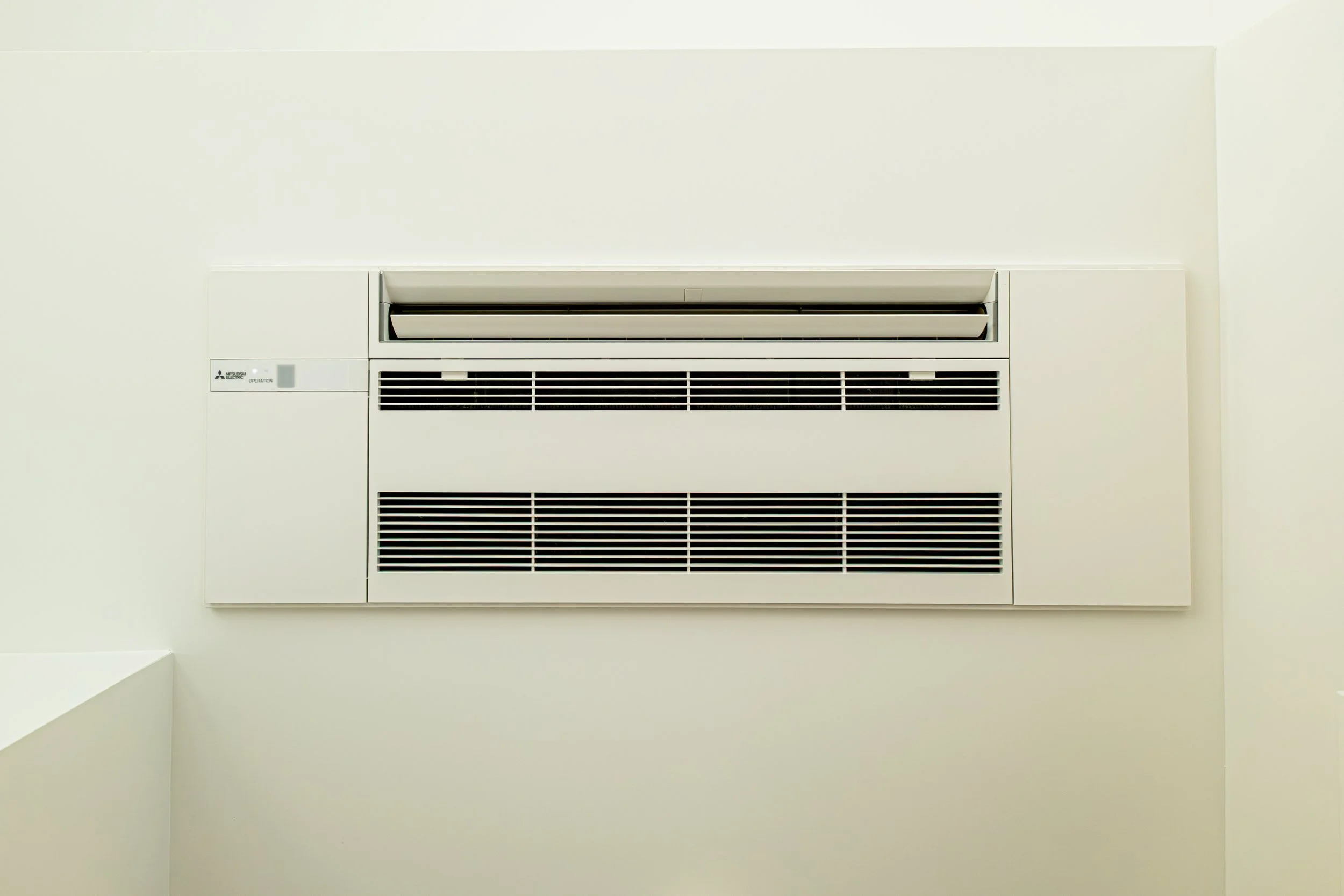 A white wall-mounted air conditioning unit with black vents, set against a plain beige wall.