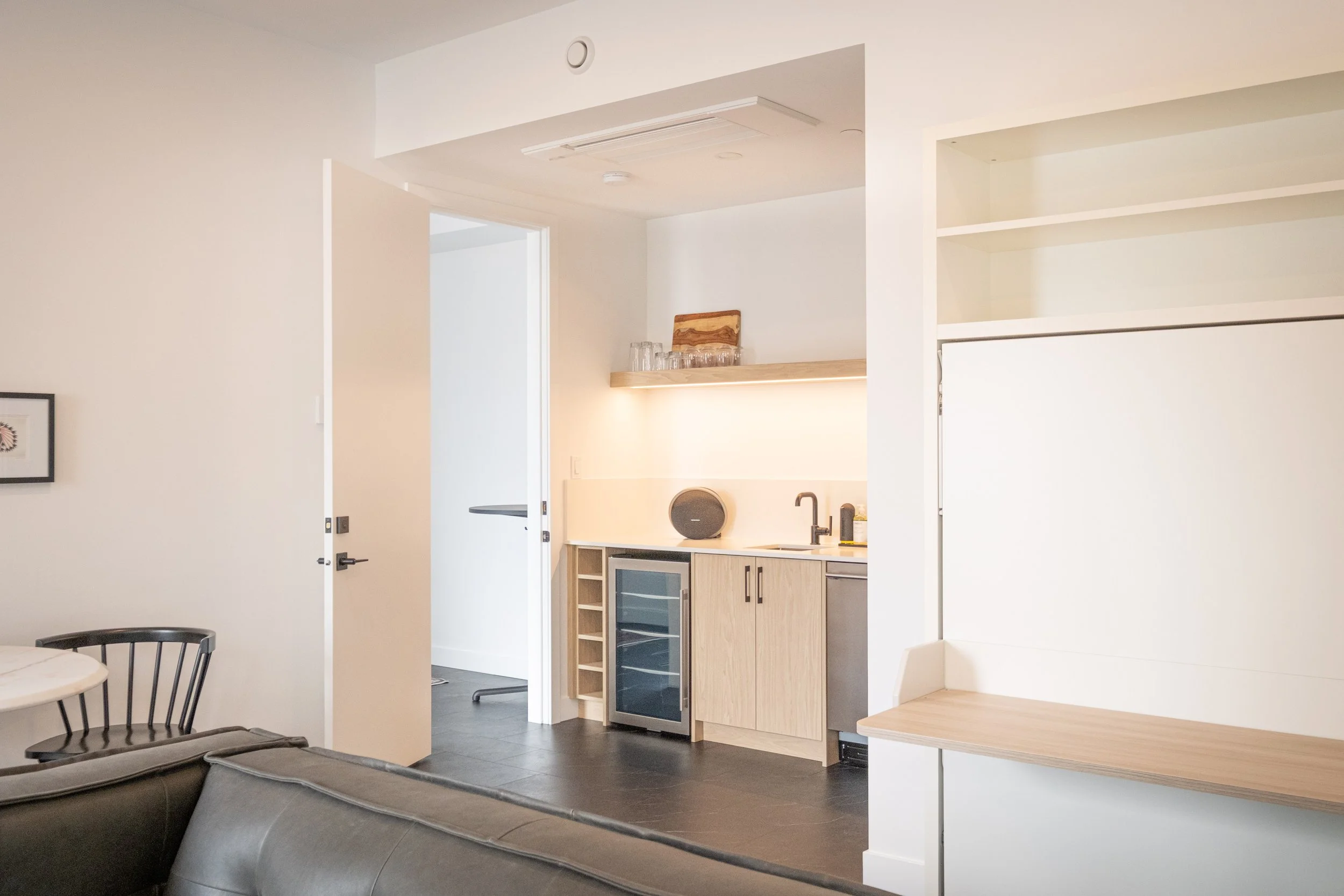 Interior view of a modern apartment with a kitchenette, black leather sofa, chairs, and white walls.