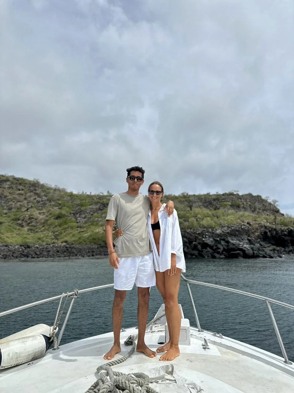A young man and woman standing on the bow of a boat, with the man in a beige t-shirt and white shorts, and the woman in a white shirt and black bikini top, both wearing sunglasses, smiling, with a green hilly landscape and cloudy sky in the background.