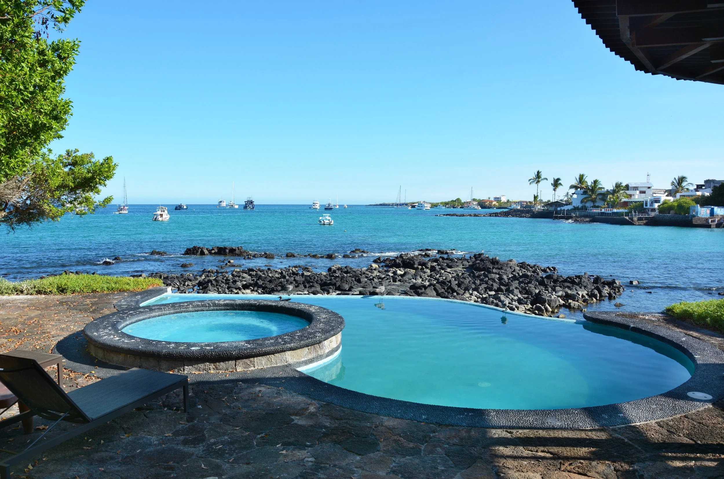 Hot tub and swimming pool overlooking a harbor with boats, rocks, and a distant shoreline with houses and palm trees on a sunny day.