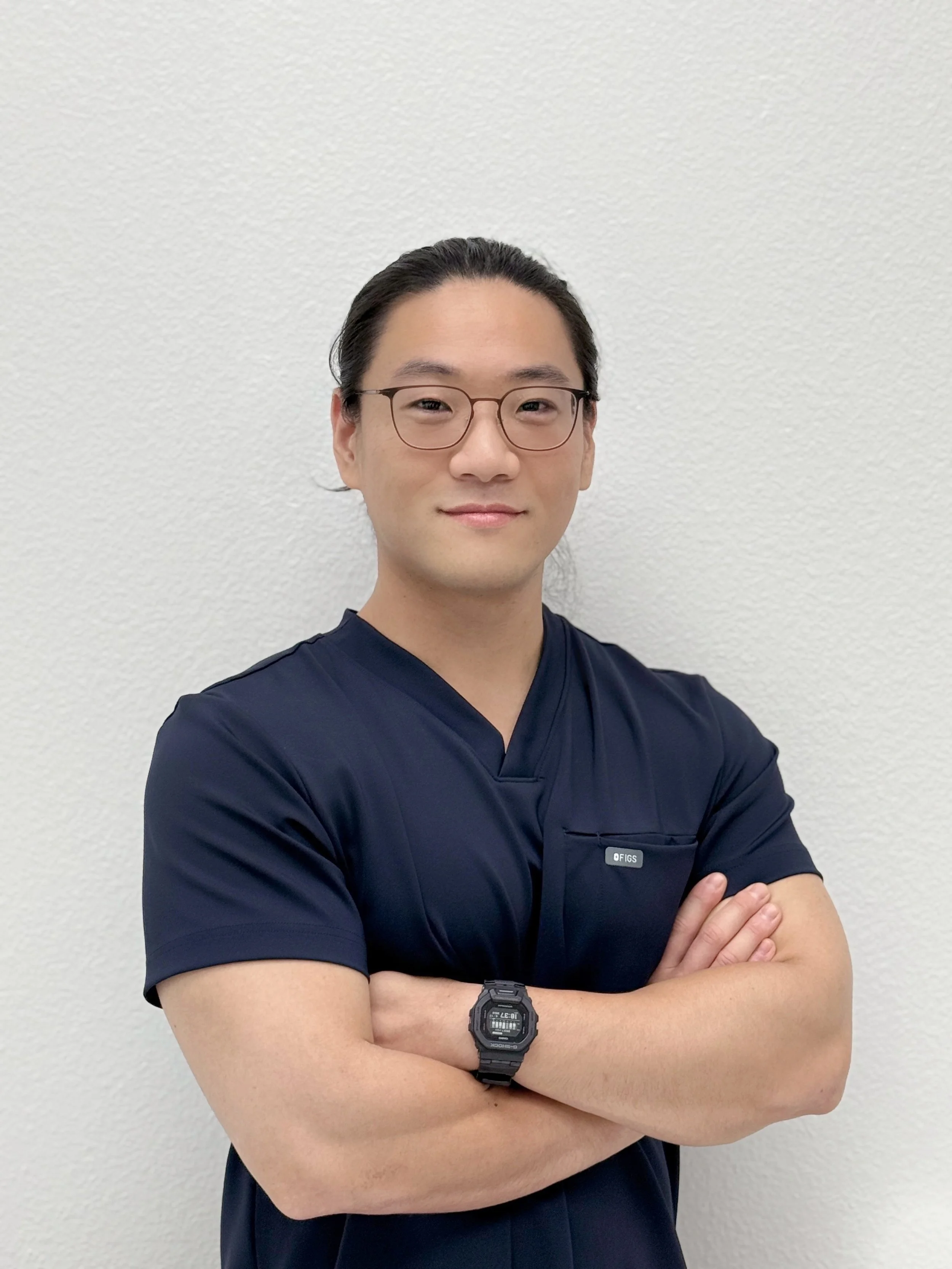 A confident man with long dark hair tied back, wearing glasses and a navy medical scrub top, standing against a plain white wall with arms crossed and smiling gently.