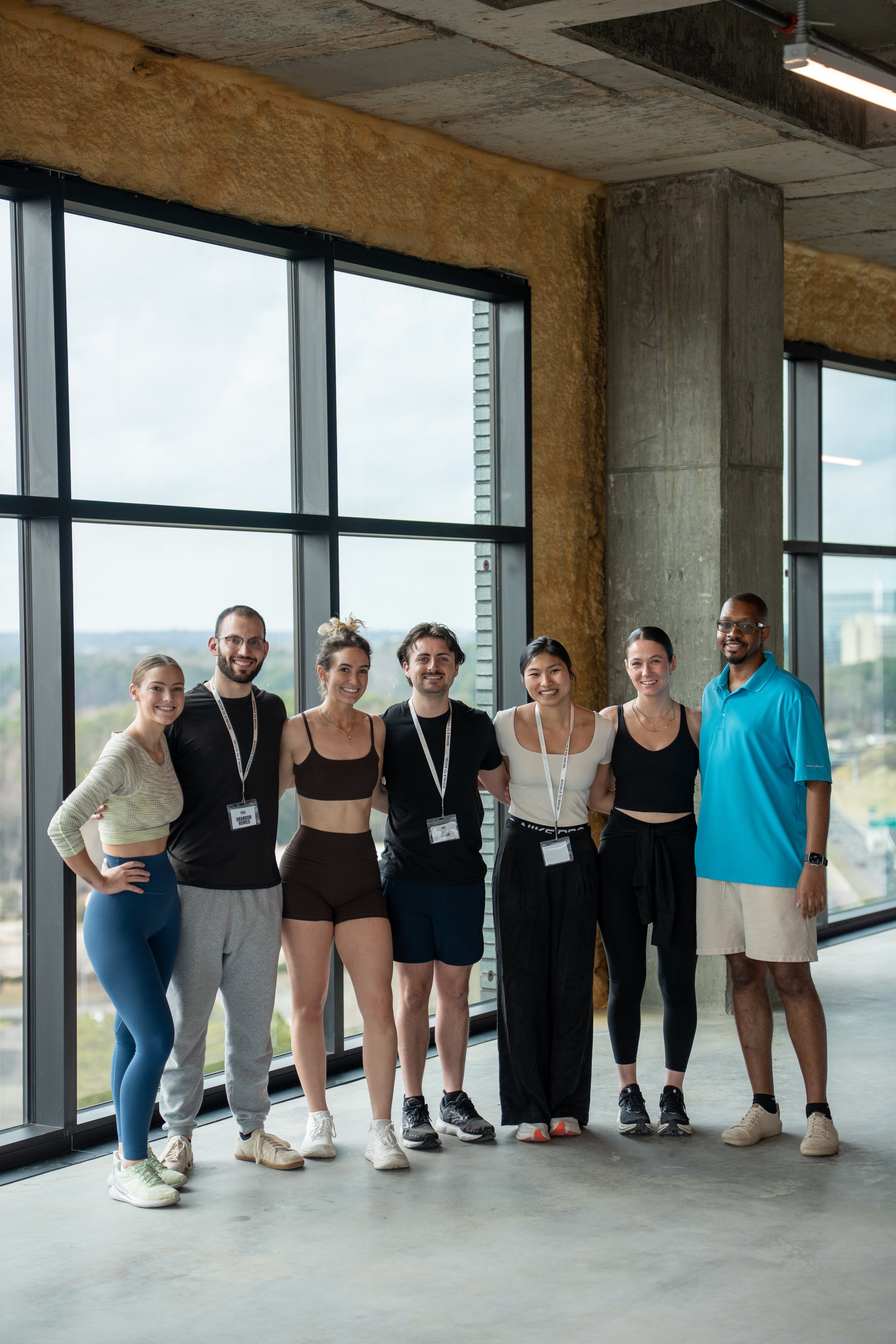 Group of seven diverse people in casual athletic clothing standing in a modern building with large windows showing a city view, smiling at the camera.