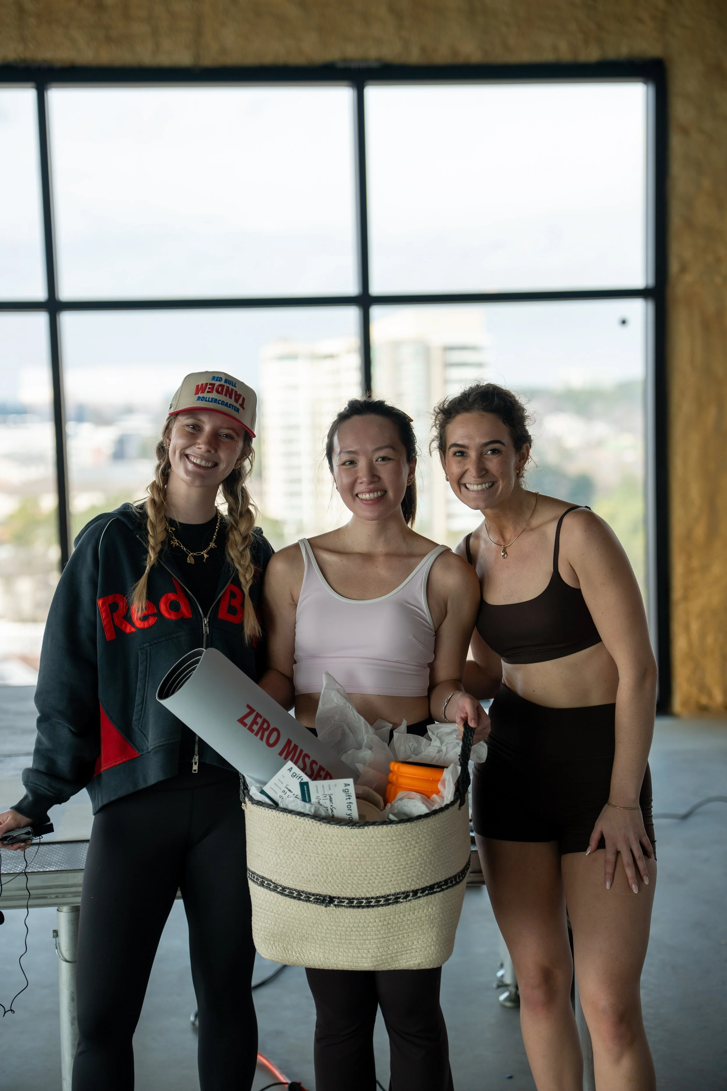 Three women smiling, standing together indoors, holding a basket with items, in front of a large window.