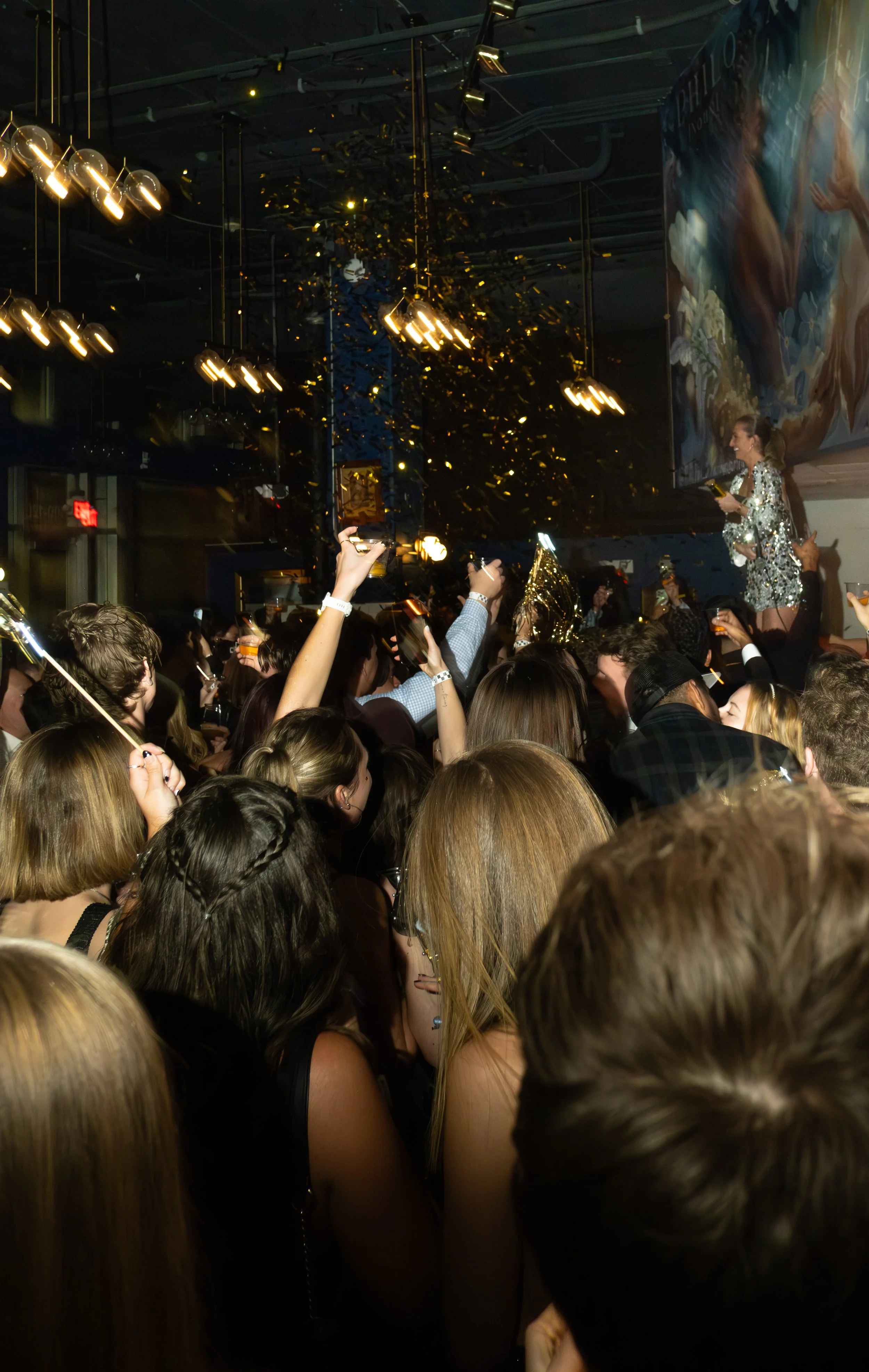 Crowd of people celebrating at a party or concert, with some holding drinks and wearing party hats, in a dimly lit venue with hanging lights and confetti falling from above.