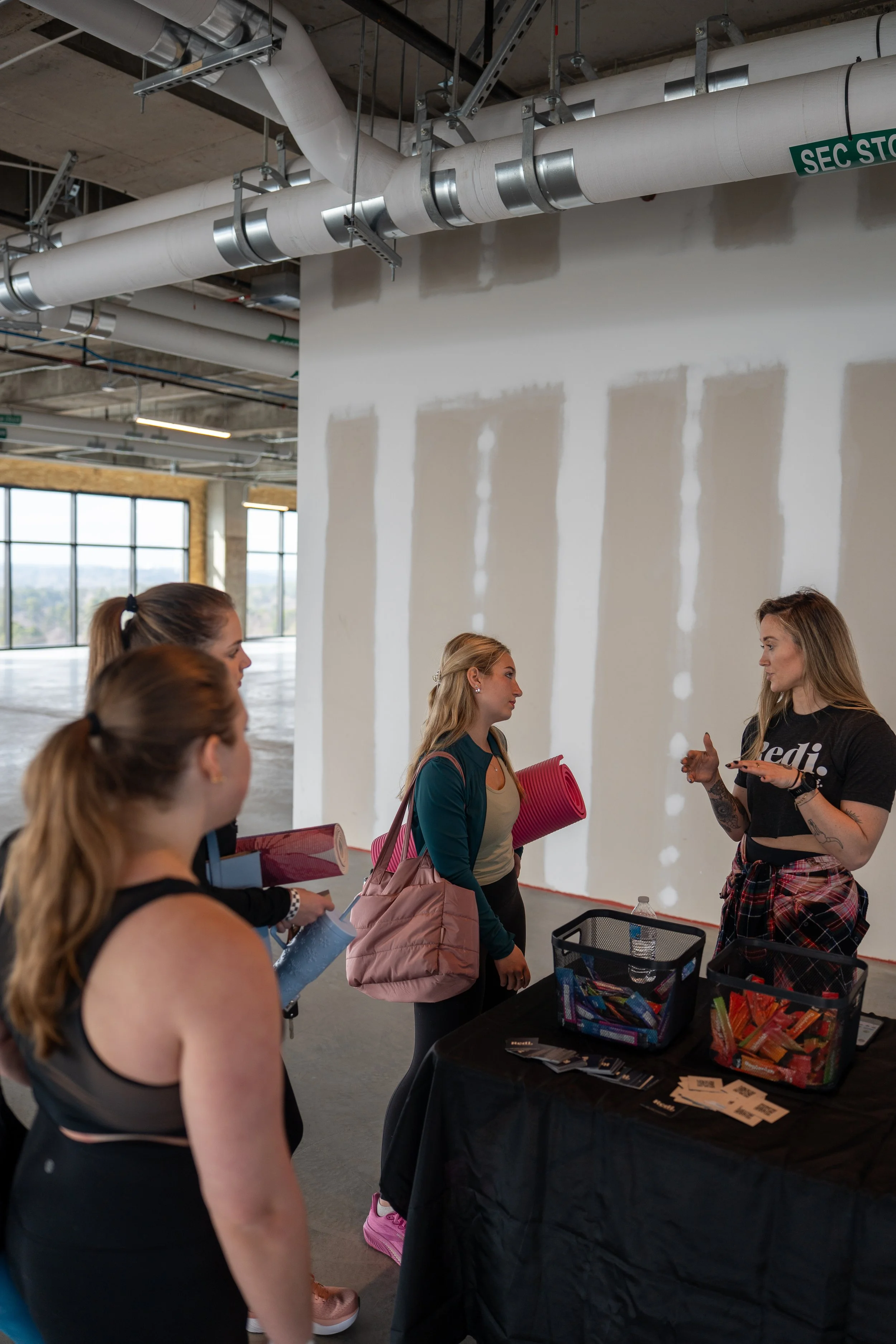 A group of women in workout attire listening to a woman in a black t-shirt with white text, explaining something at a table with snacks and flyers inside an unfinished building with exposed ducts and drywall.