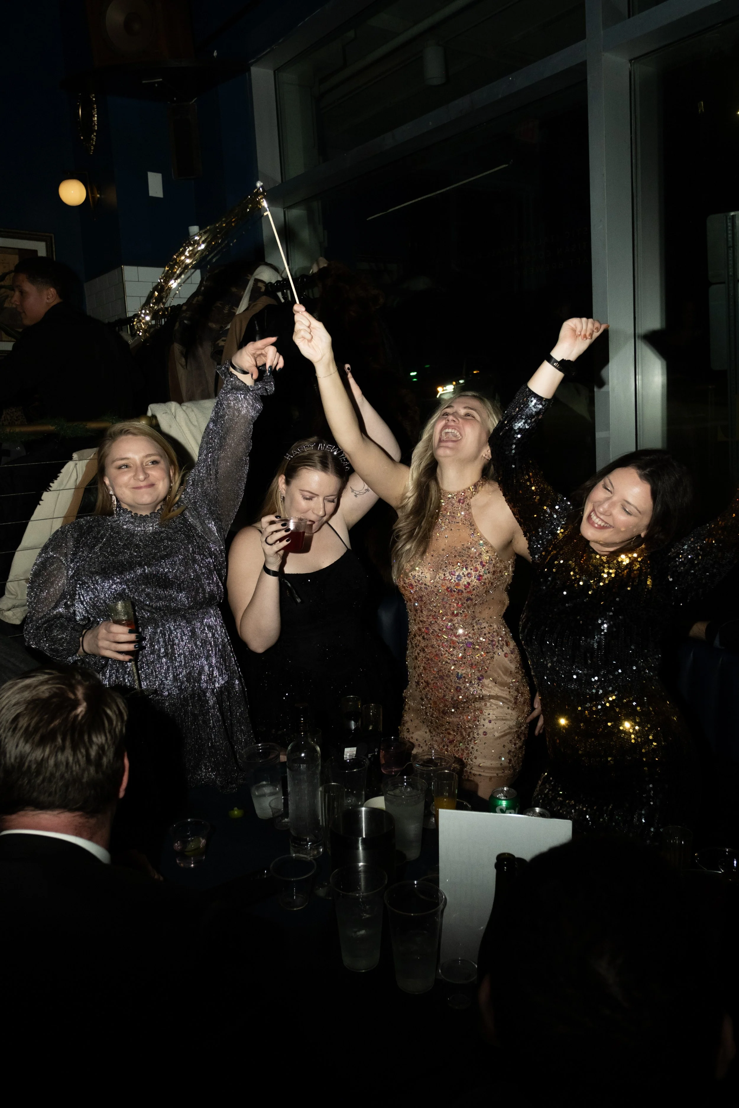 Group of women celebrating at a party, wearing sparkly dresses, with drinks in their hands, dancing and smiling.