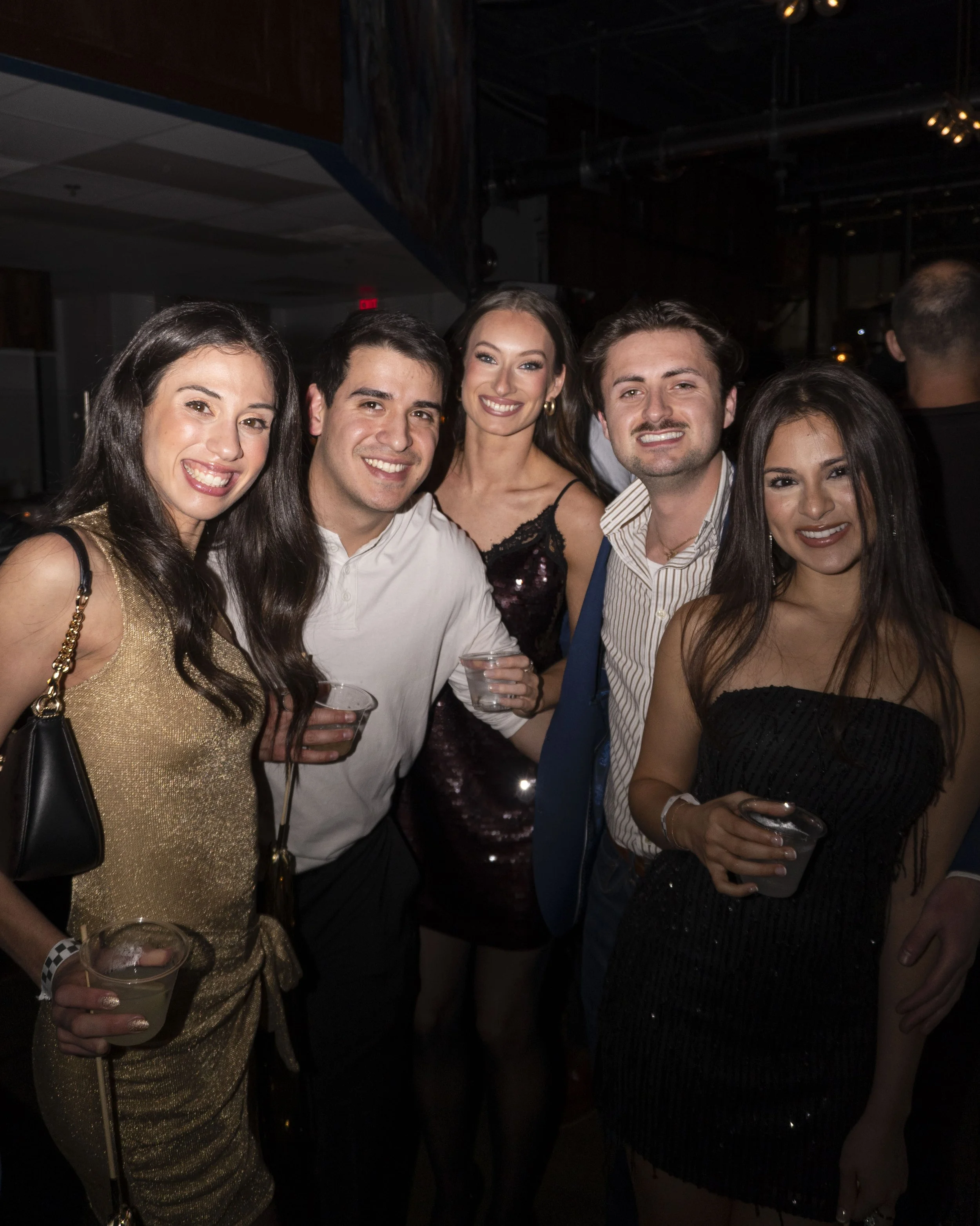 Group of five friends at a party, smiling and holding drinks, dressed in evening attire.