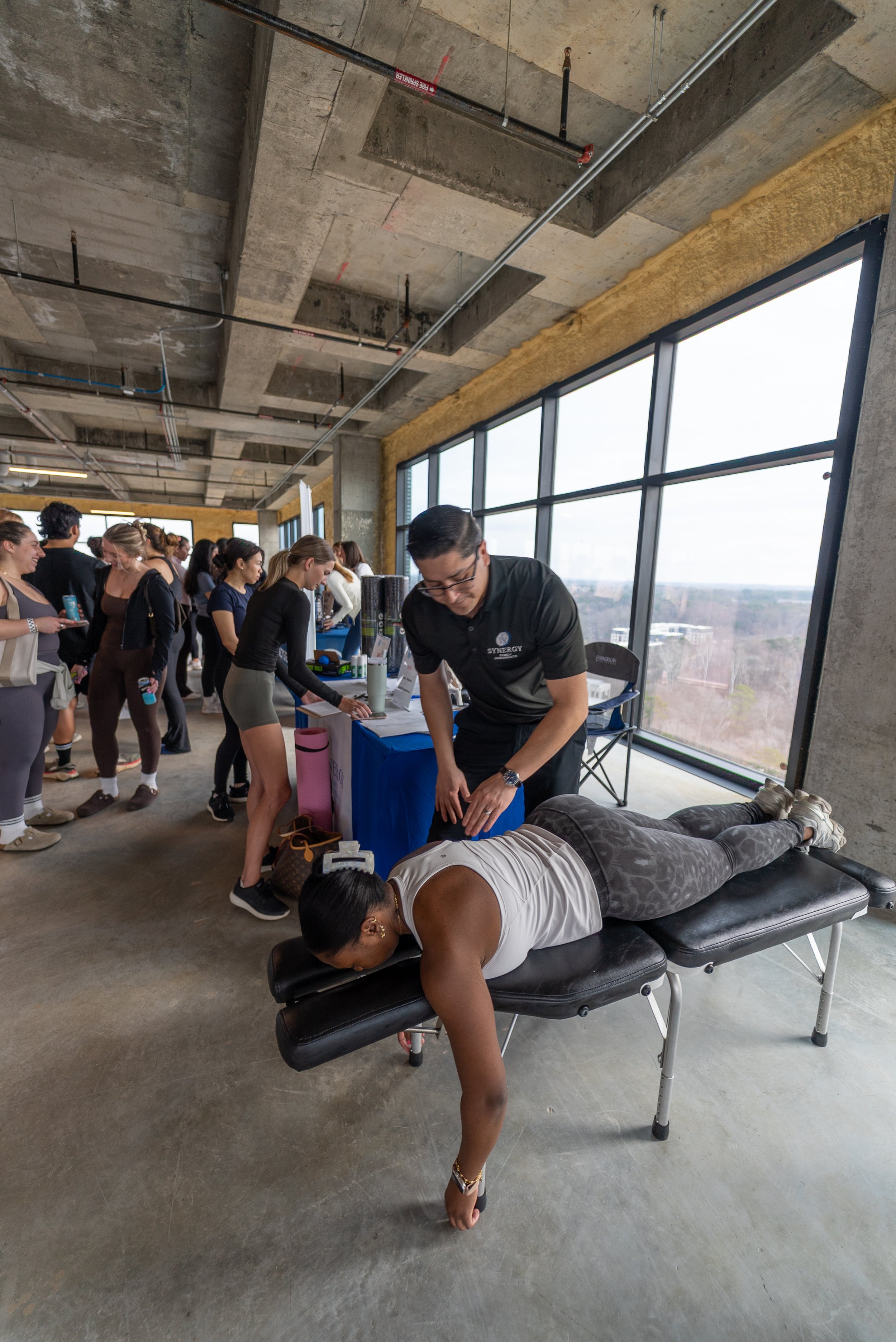 A person receiving a massage on a massage table in a spacious, industrial-style room with large windows and a crowd of people waiting in line.