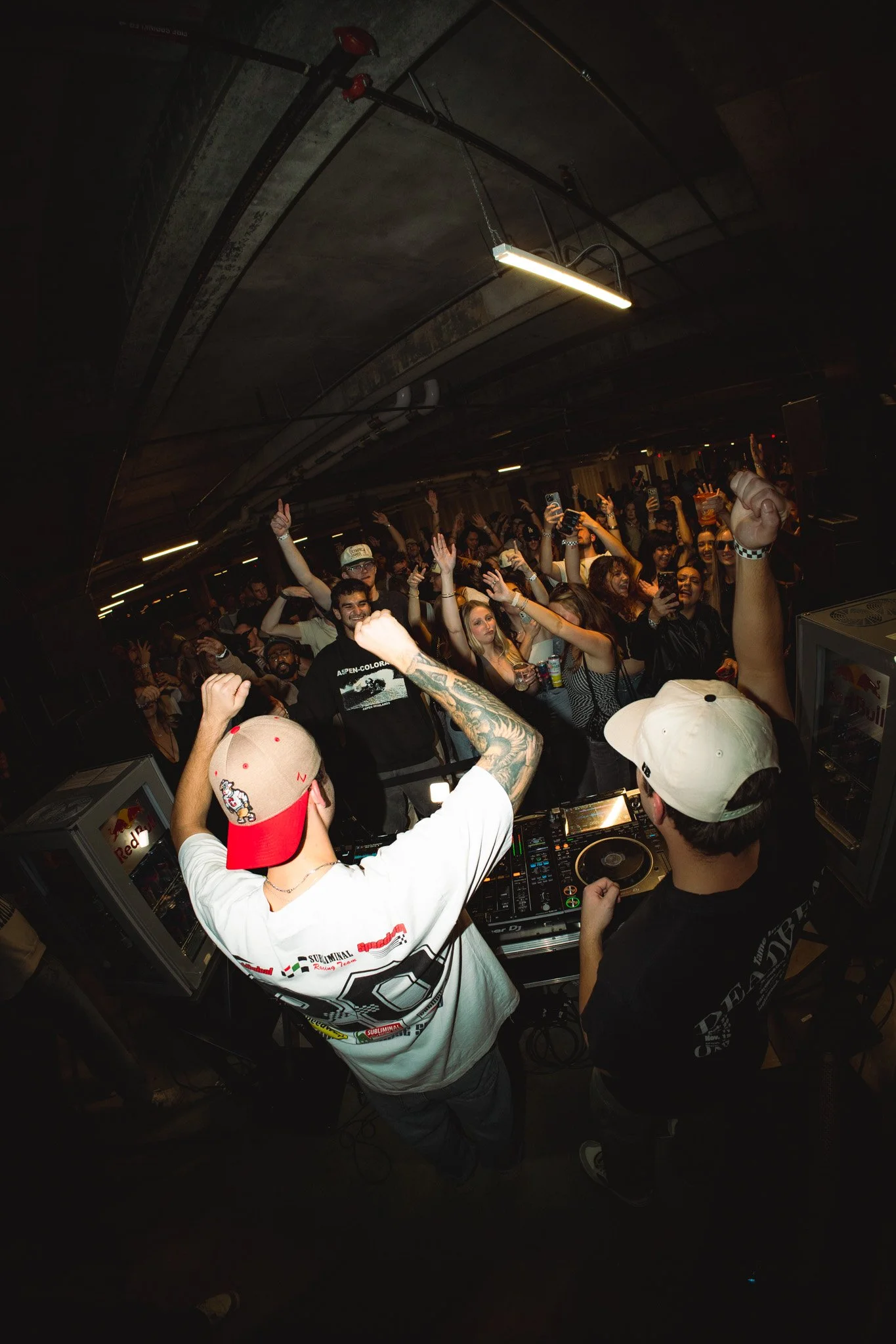 Crowd dancing at a nightclub with DJs in the foreground, raising their fists and enjoying the music.