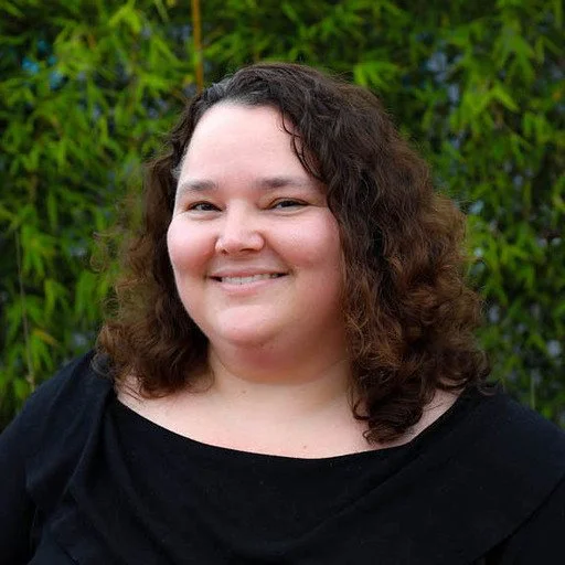 A woman with curly brown hair smiling outdoors with green foliage in the background.