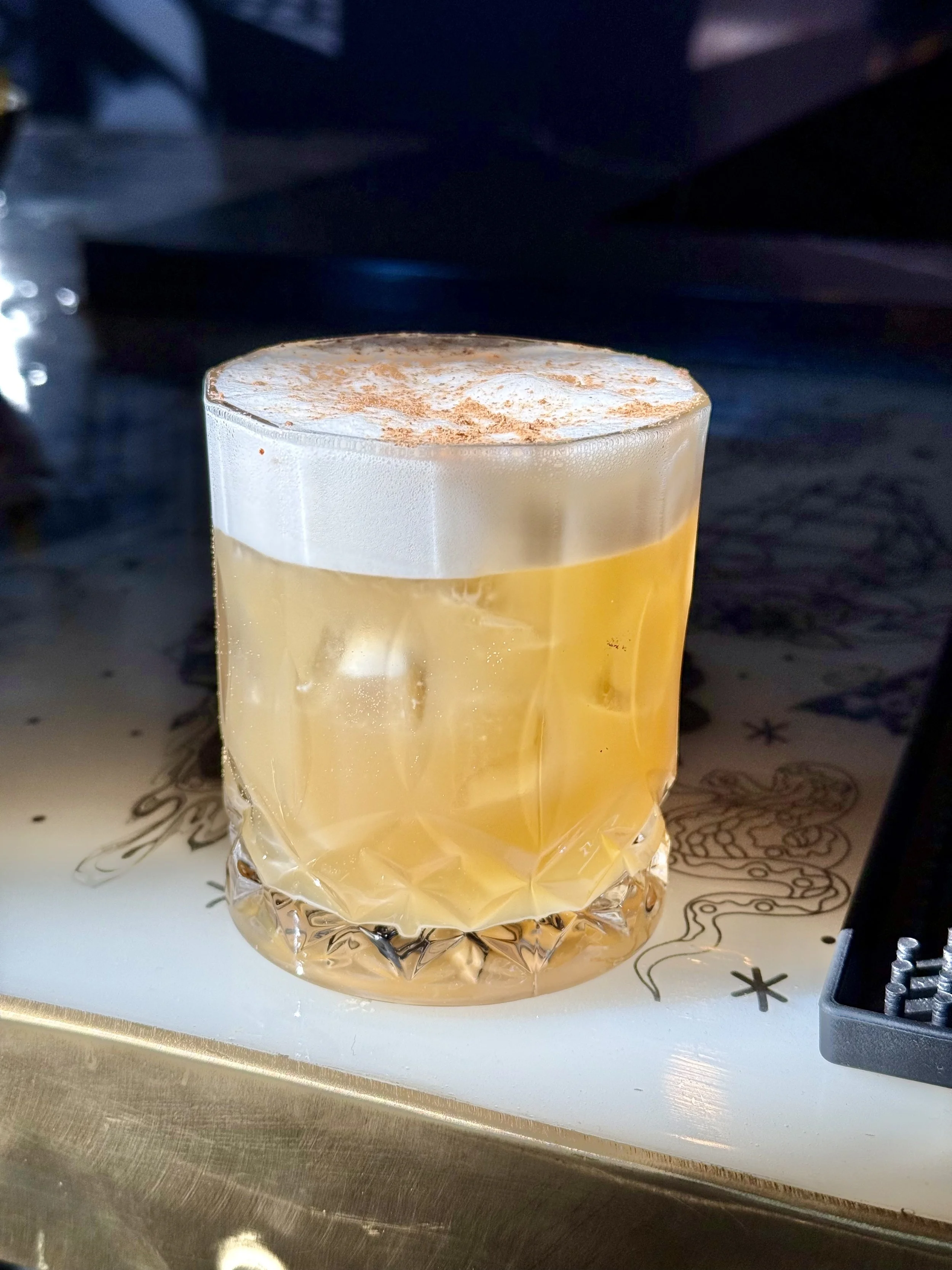 A cocktail with a thick layer of foamy topping in a textured glass on a decorated surface.