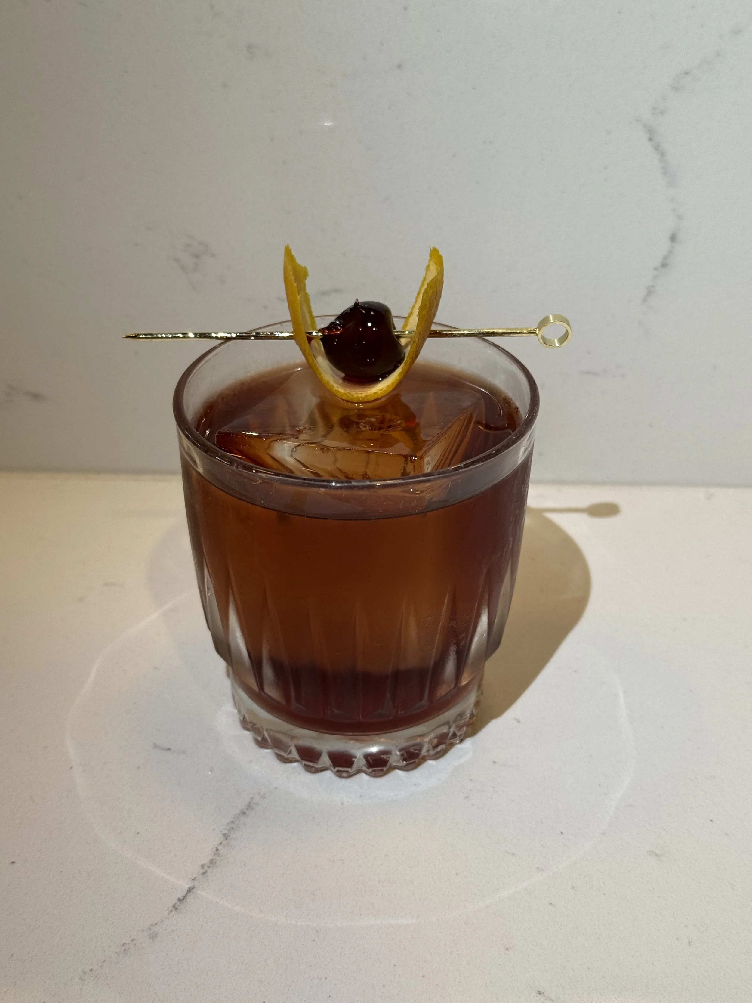 A glass of dark cocktail with ice, garnished with a lemon peel and a cherry on a toothpick.
