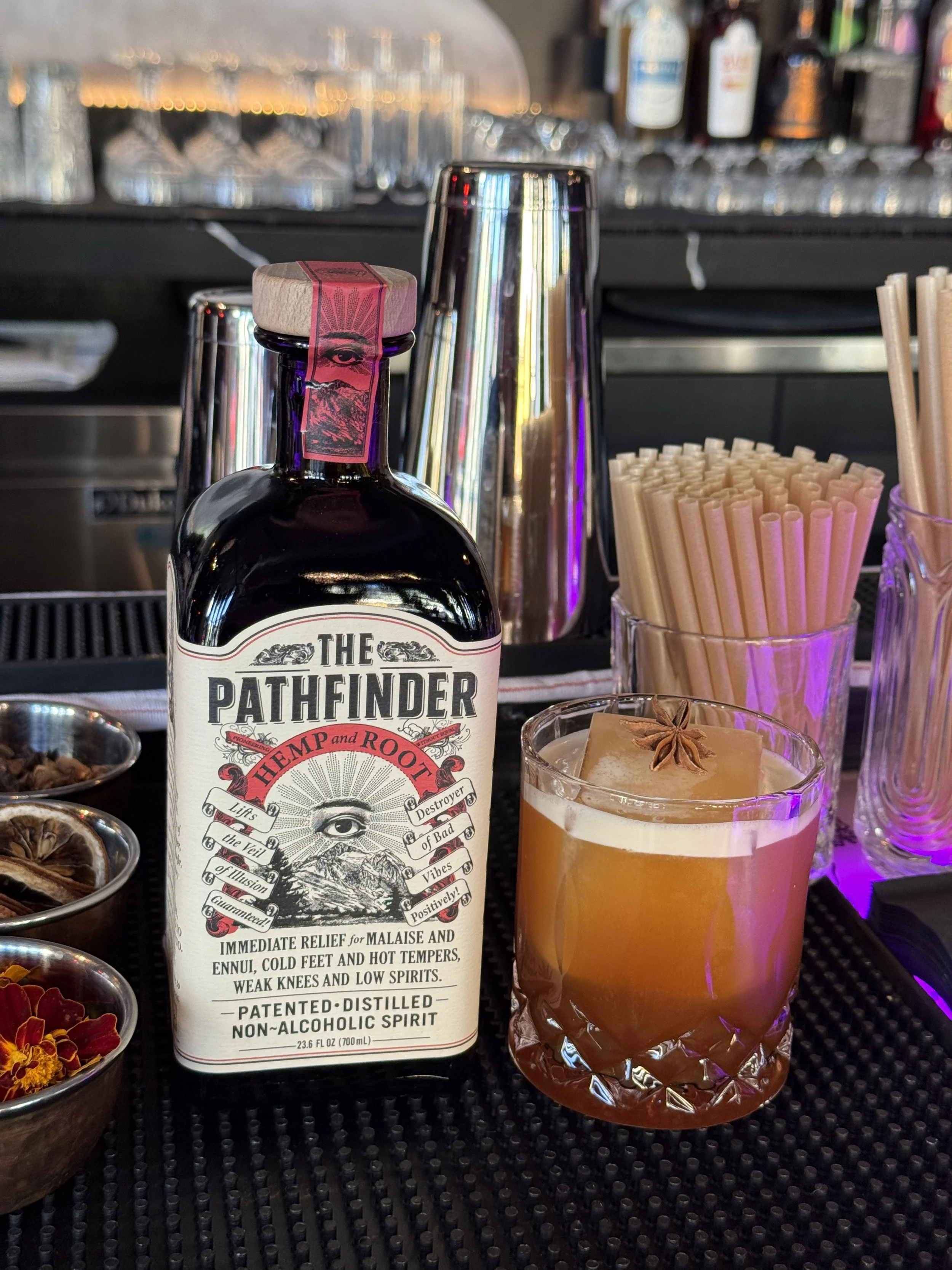 Bottle of The Pathfinder Hemp and Root non-alcoholic spirit with a cocktail garnished with a star anise in a glass, placed on a bar counter with bar tools and glasses in the background.