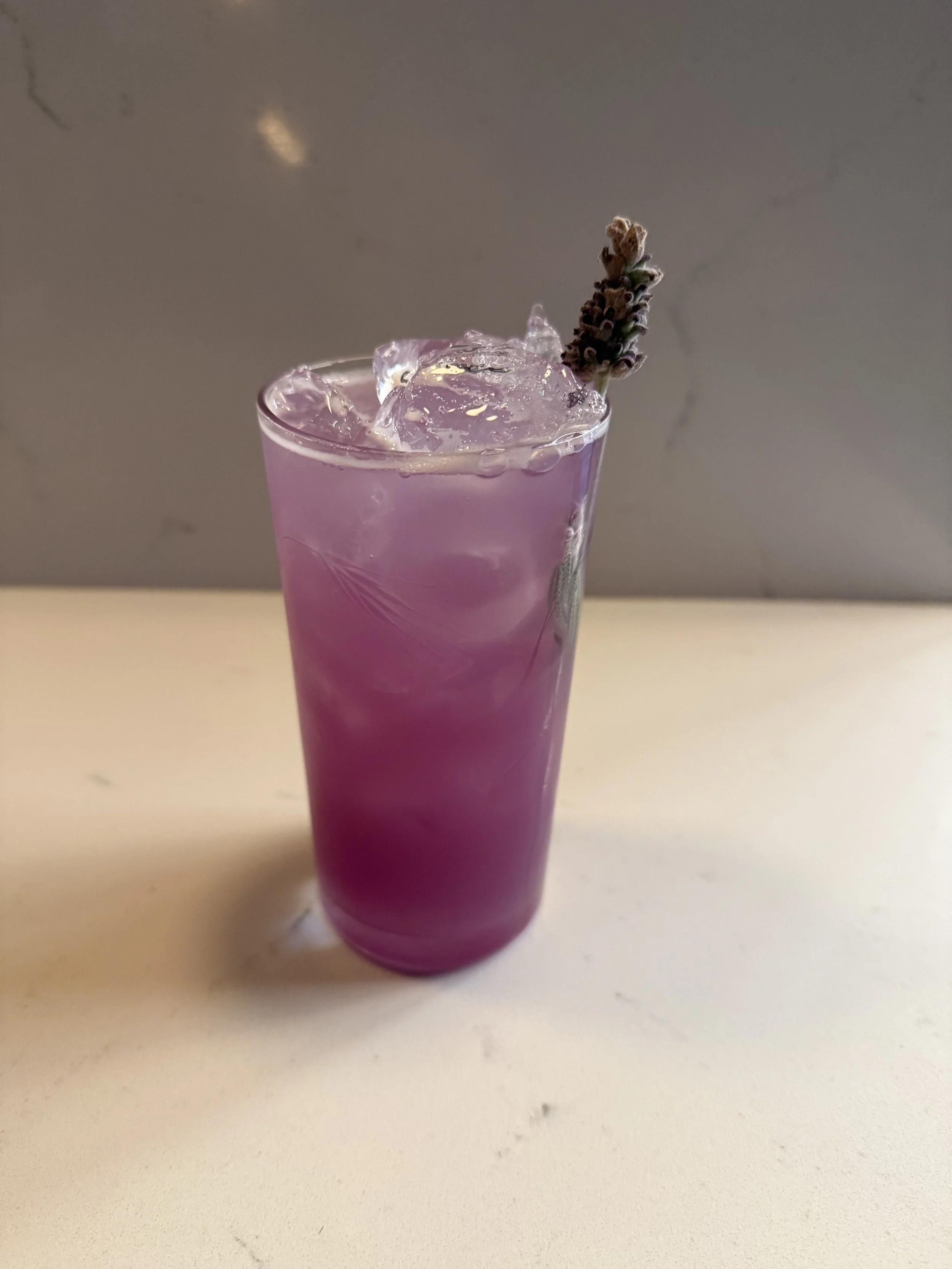A tall glass of purple lavender-colored beverage with ice cubes and a sprig of lavender as garnish on a beige surface.