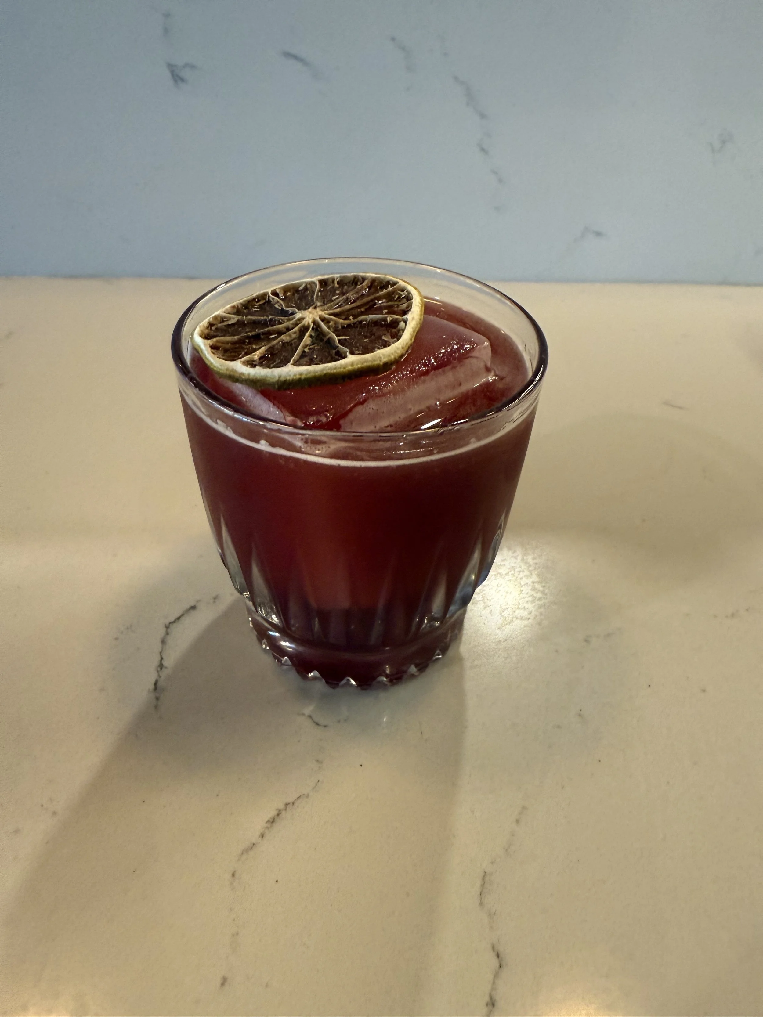A glass of a dark red beverage with a slice of dried lemon on top, served on a light-colored marble countertop.