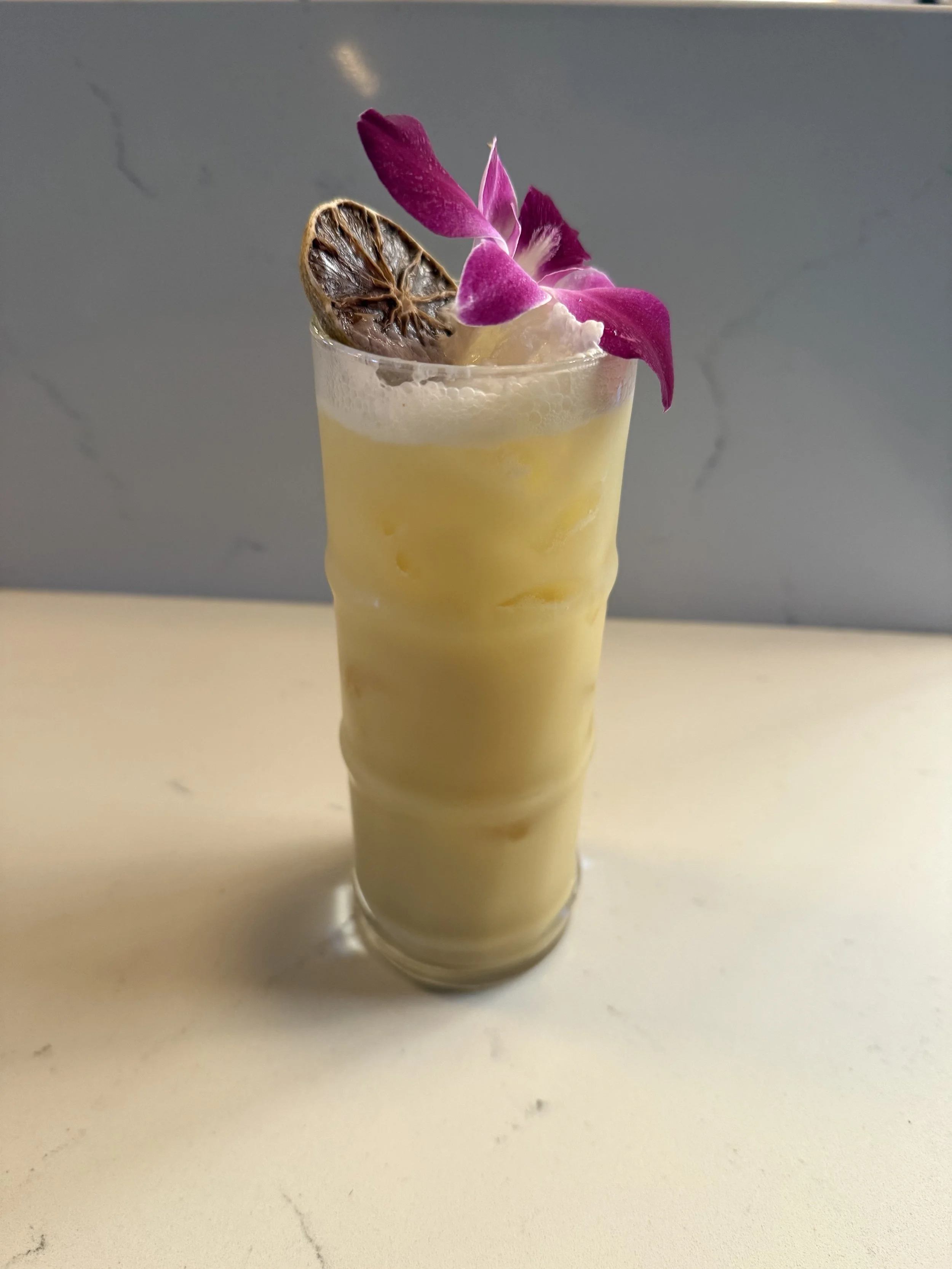 A tall glass of yellow beverage garnished with a dried lime slice and purple orchid flowers placed on top.