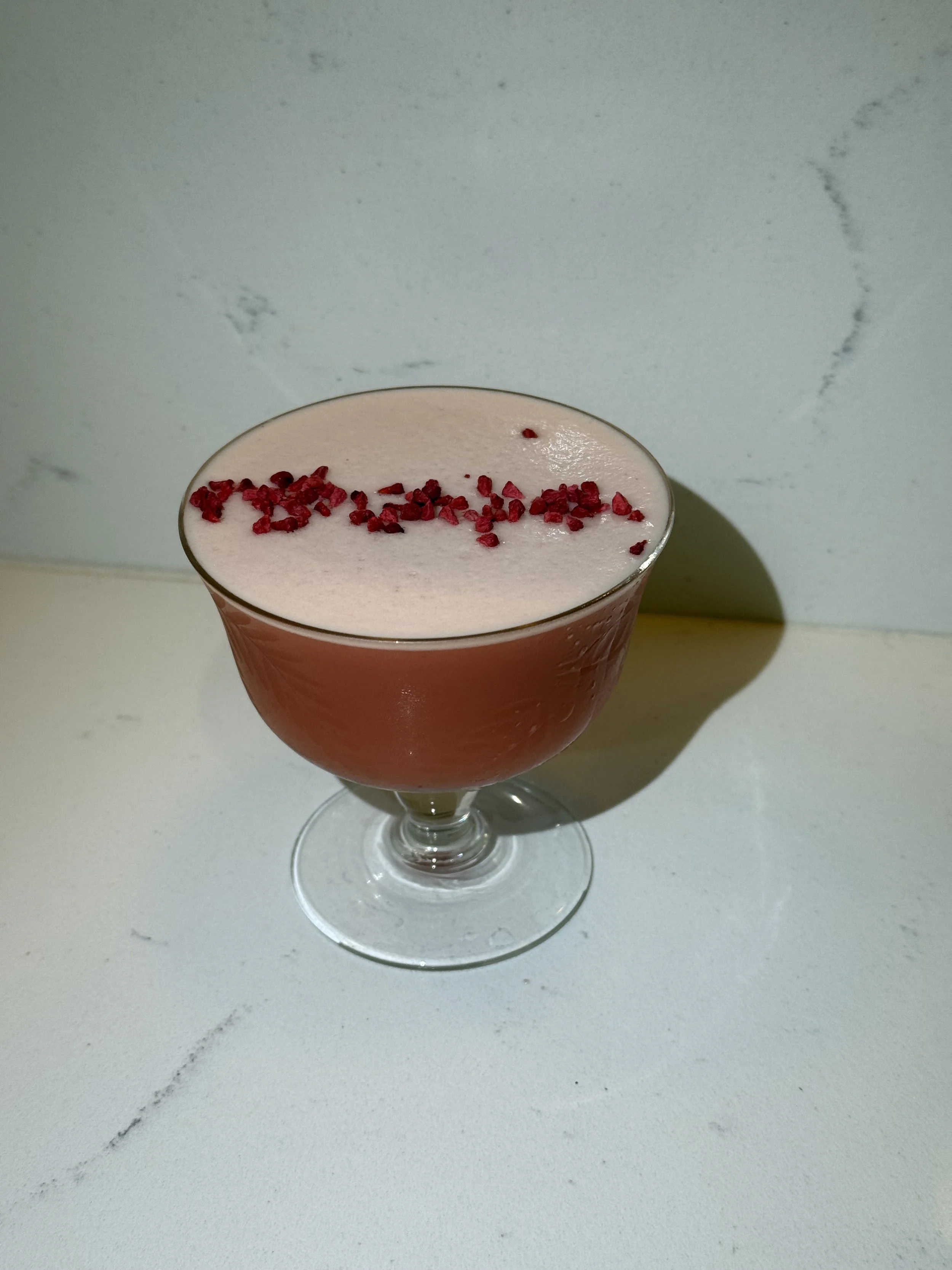 A pink cocktail garnished with red crushed candies in a stemmed glass, placed on a white surface with a light background.