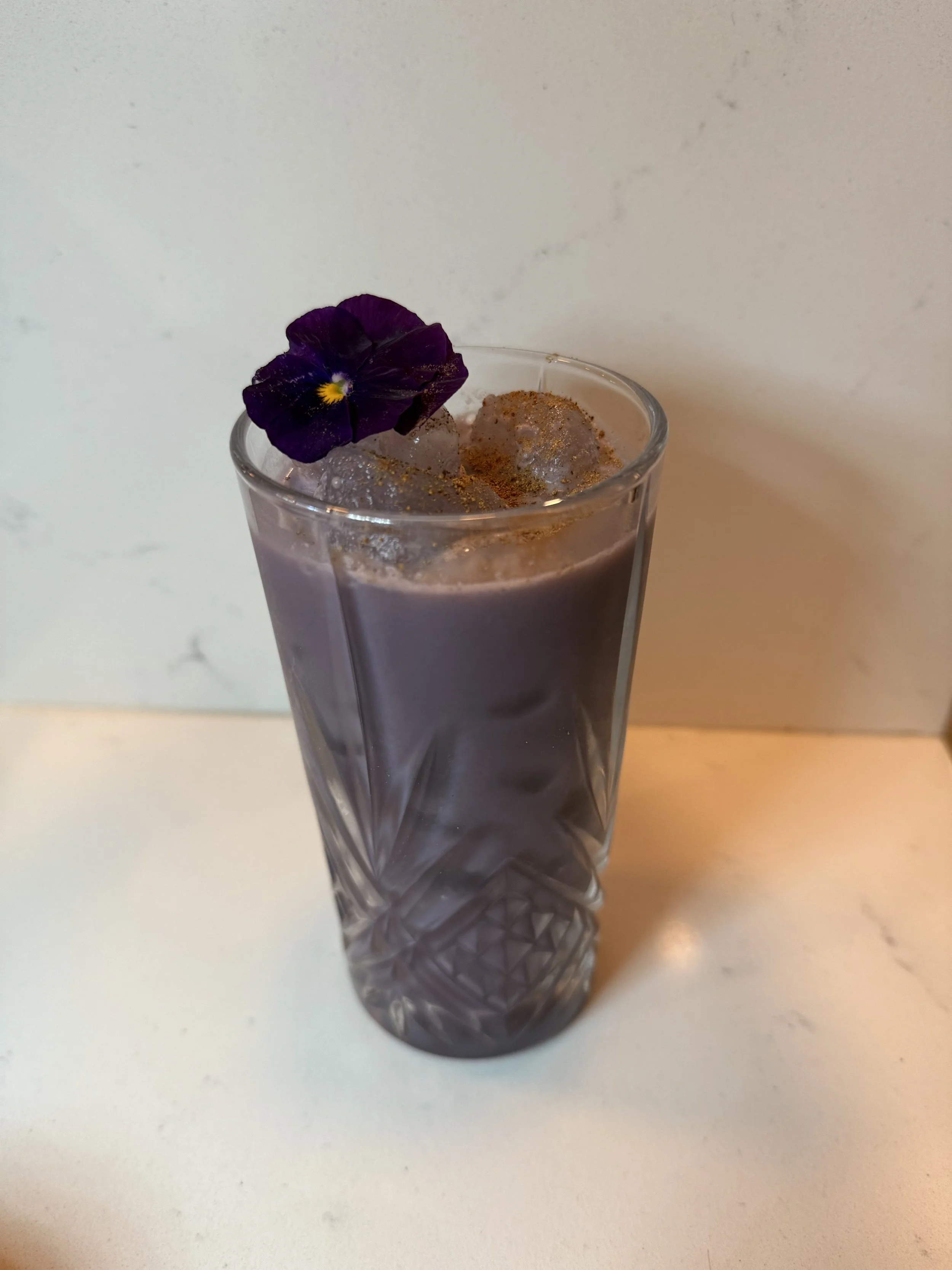A tall glass of purple-colored beverage garnished with a purple pansy flower on ice and sprinkled with a brown powder, set against a plain light background.