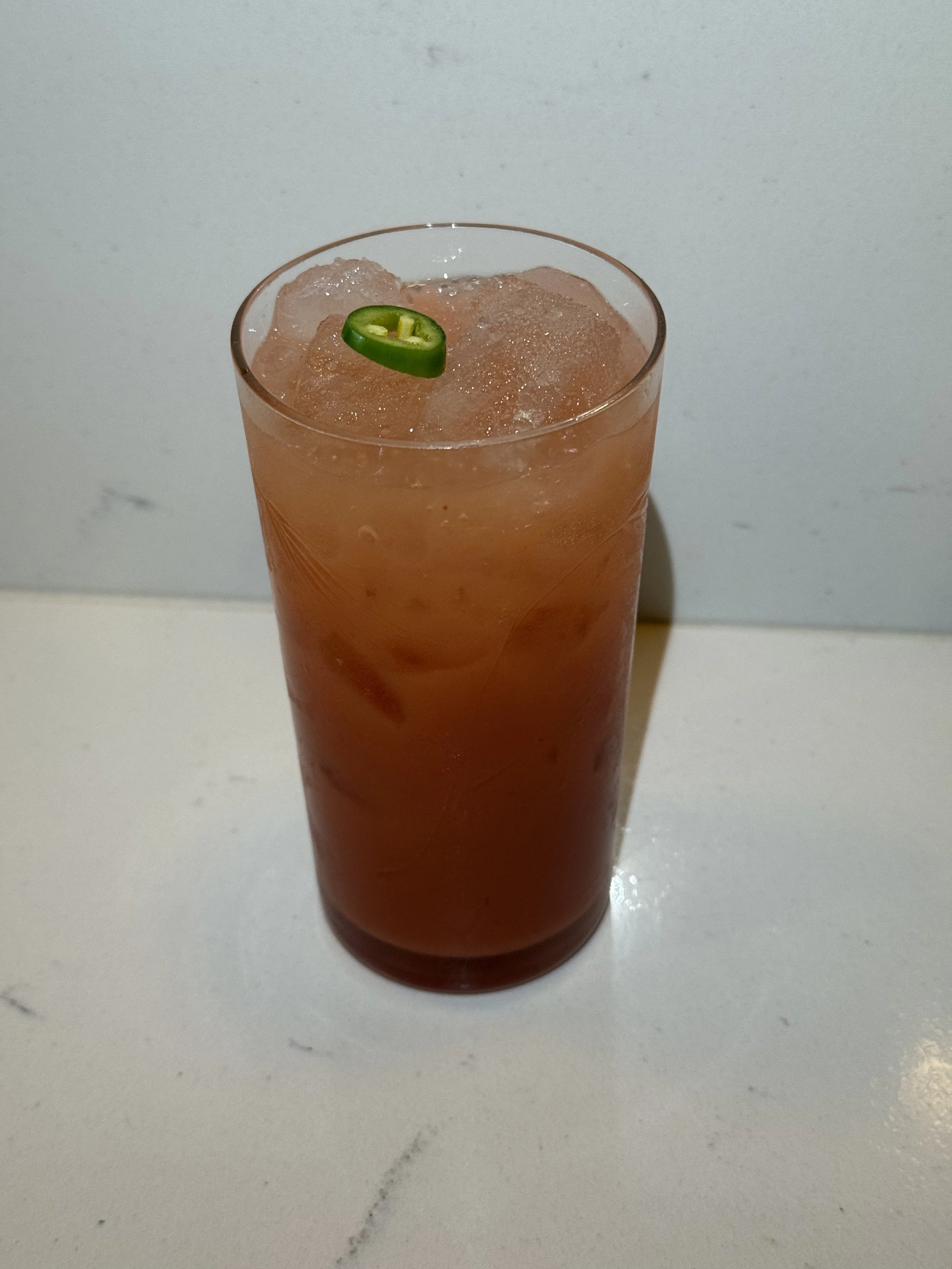 A tall glass of iced tea garnished with a slice of jalapeño pepper on top, placed on a white surface with a plain background.