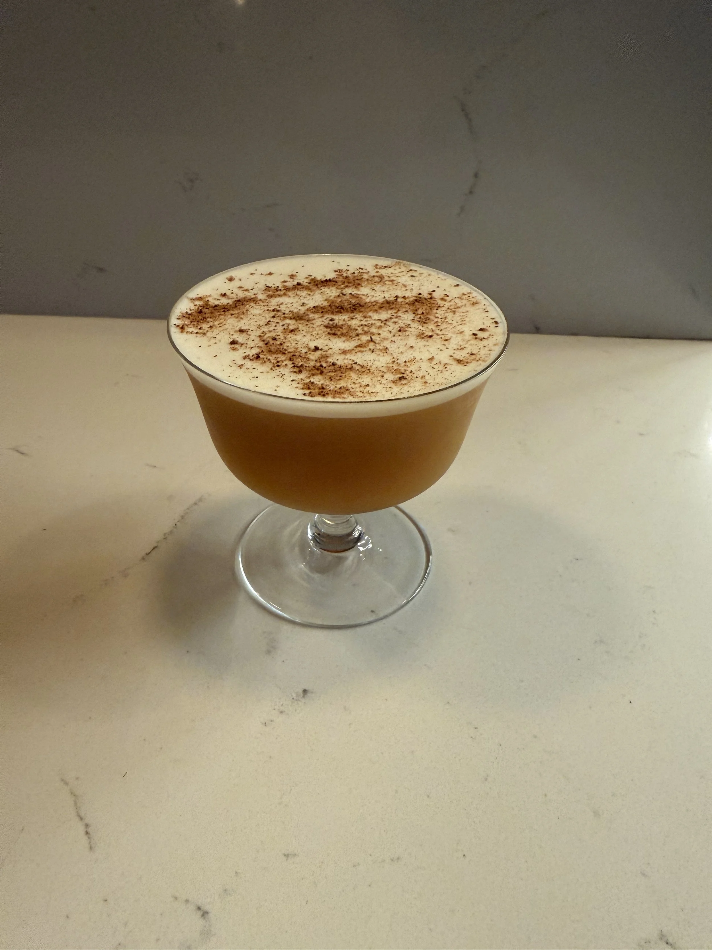 A cocktail in a clear glass with a wide bowl and short stem, topped with whipped cream and sprinkled with cinnamon or cocoa powder, on a white marble surface.