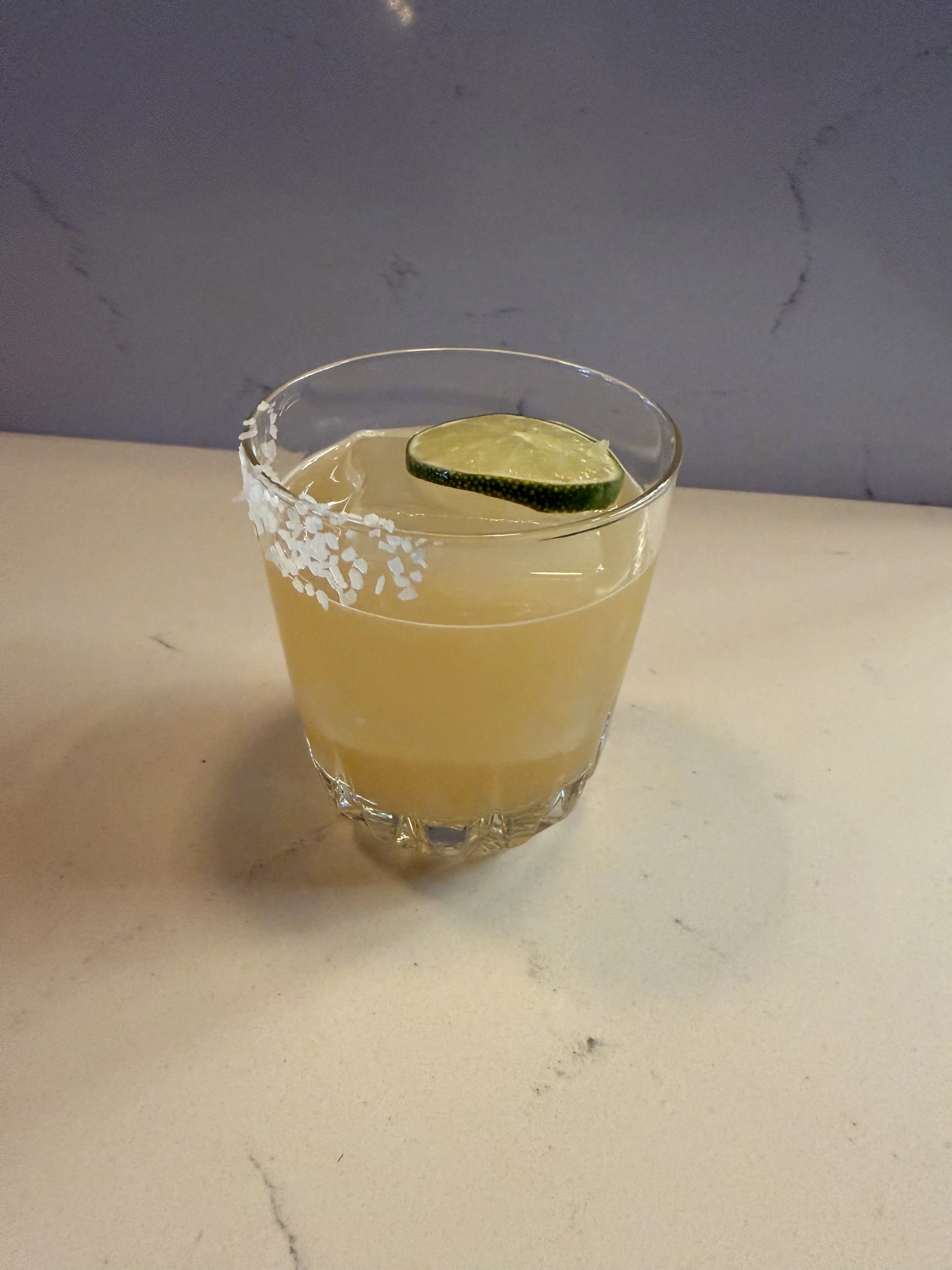 A glass of yellow cocktail with a salted rim and a lime slice garnish on top, placed on a white surface with a gray background.