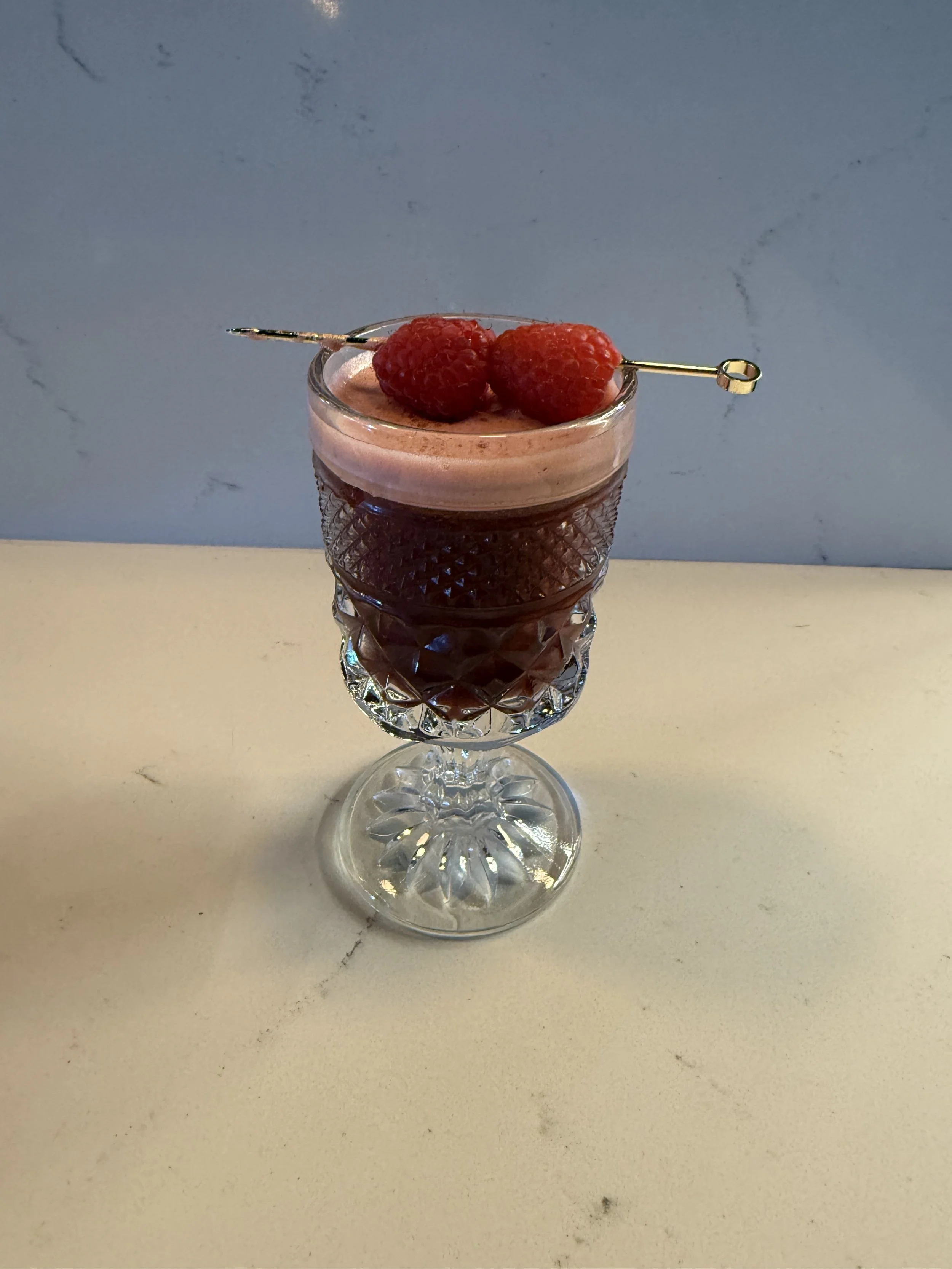 A decorative glass filled with a layered berry beverage topped with two raspberries and a cocktail pick.