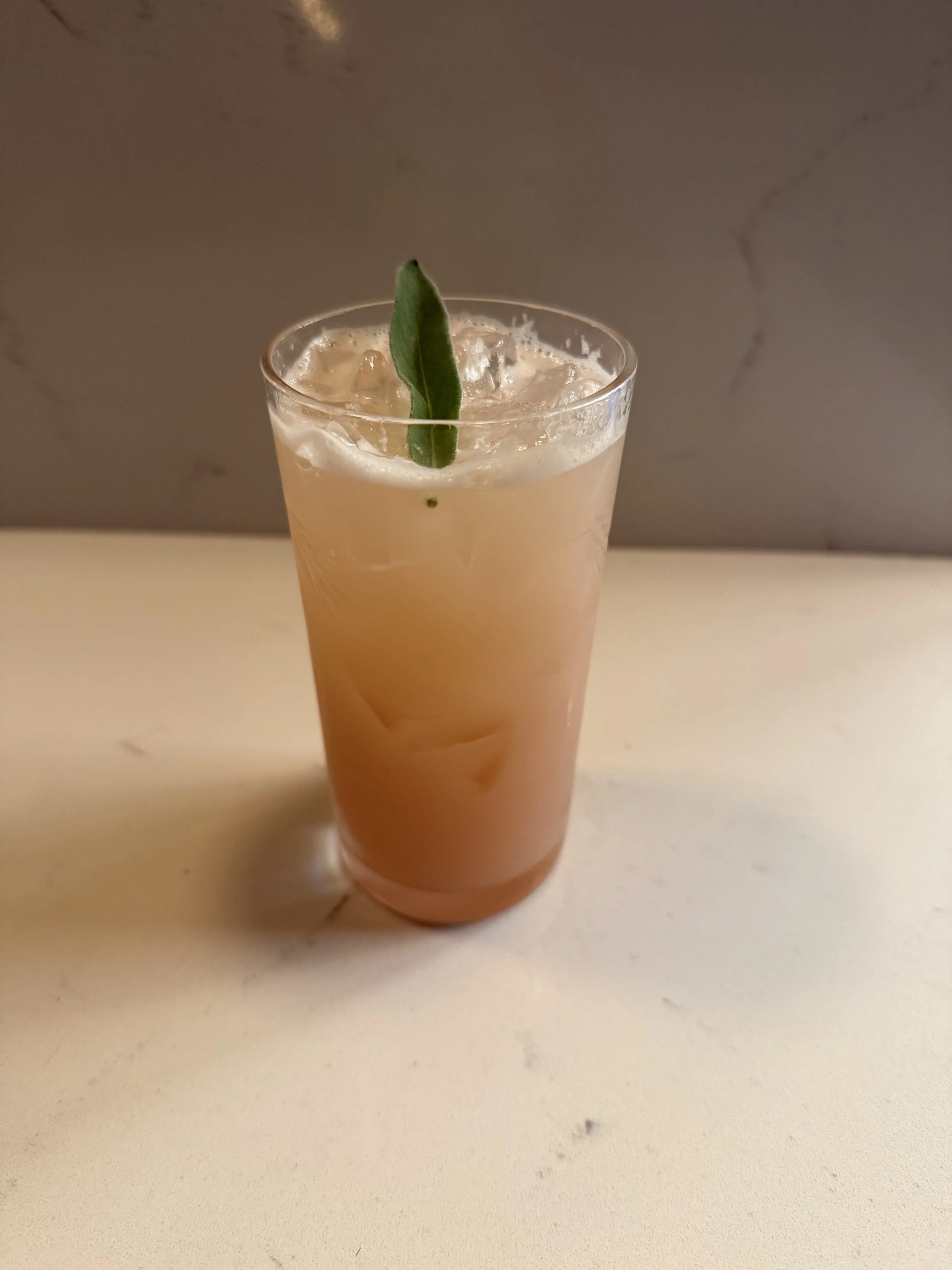 A tall glass of pink cocktail with ice and a green leaf garnish on top, placed on a light-colored surface against a gray background.