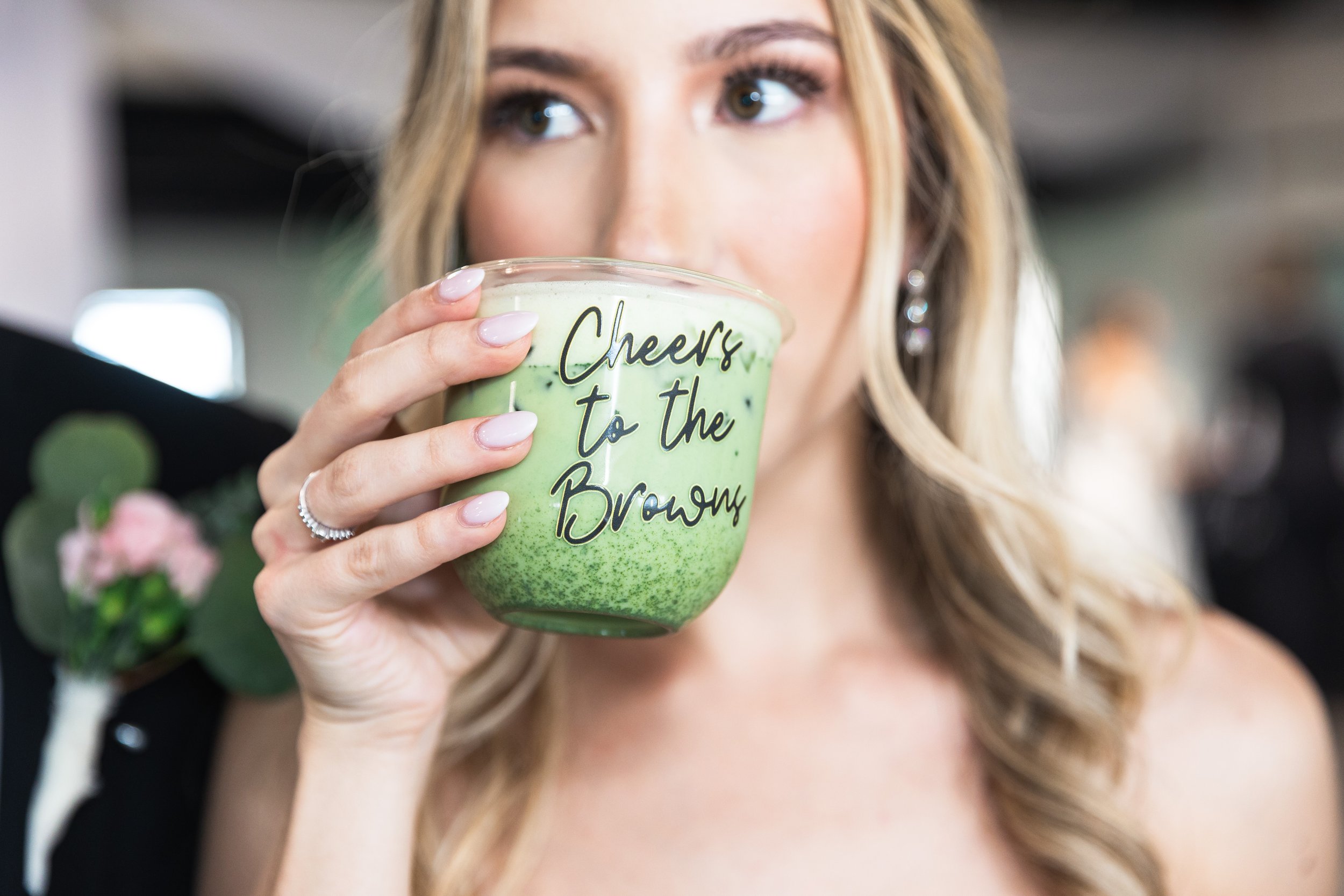 A woman with blonde hair, wearing earrings, holding a matcha latte with a straw near her mouth. The cup has the words custom cup branding, "Cheers to the Bride" written on it. Matcha Bar, Matcha Catering