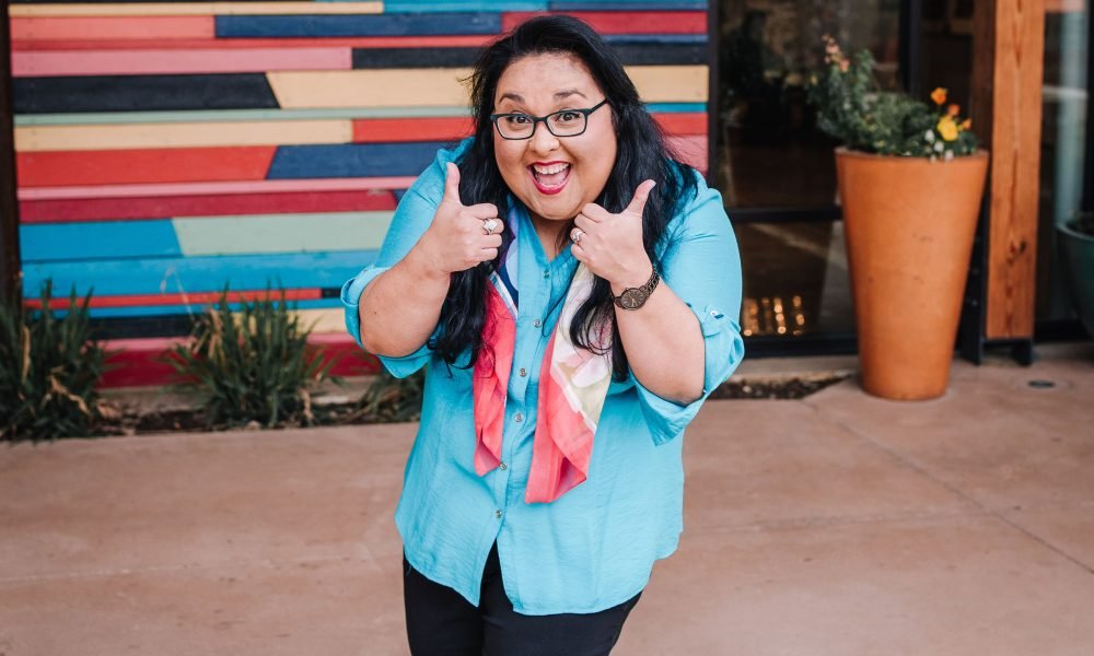 A woman with dark curly hair, glasses, and a big smile giving thumbs up outside a colorful building and potted plant.