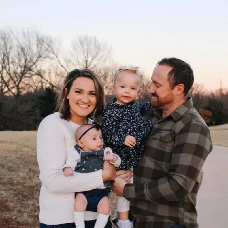 A happy family of four outdoors during sunset, with bare trees in the background. The mother, with shoulder-length brown hair, is smiling and holding a baby girl dressed in a navy dress with a headband. The father, wearing a gray checkered shirt, is smiling and holding a young girl in a navy floral dress. The family appears joyful and close.