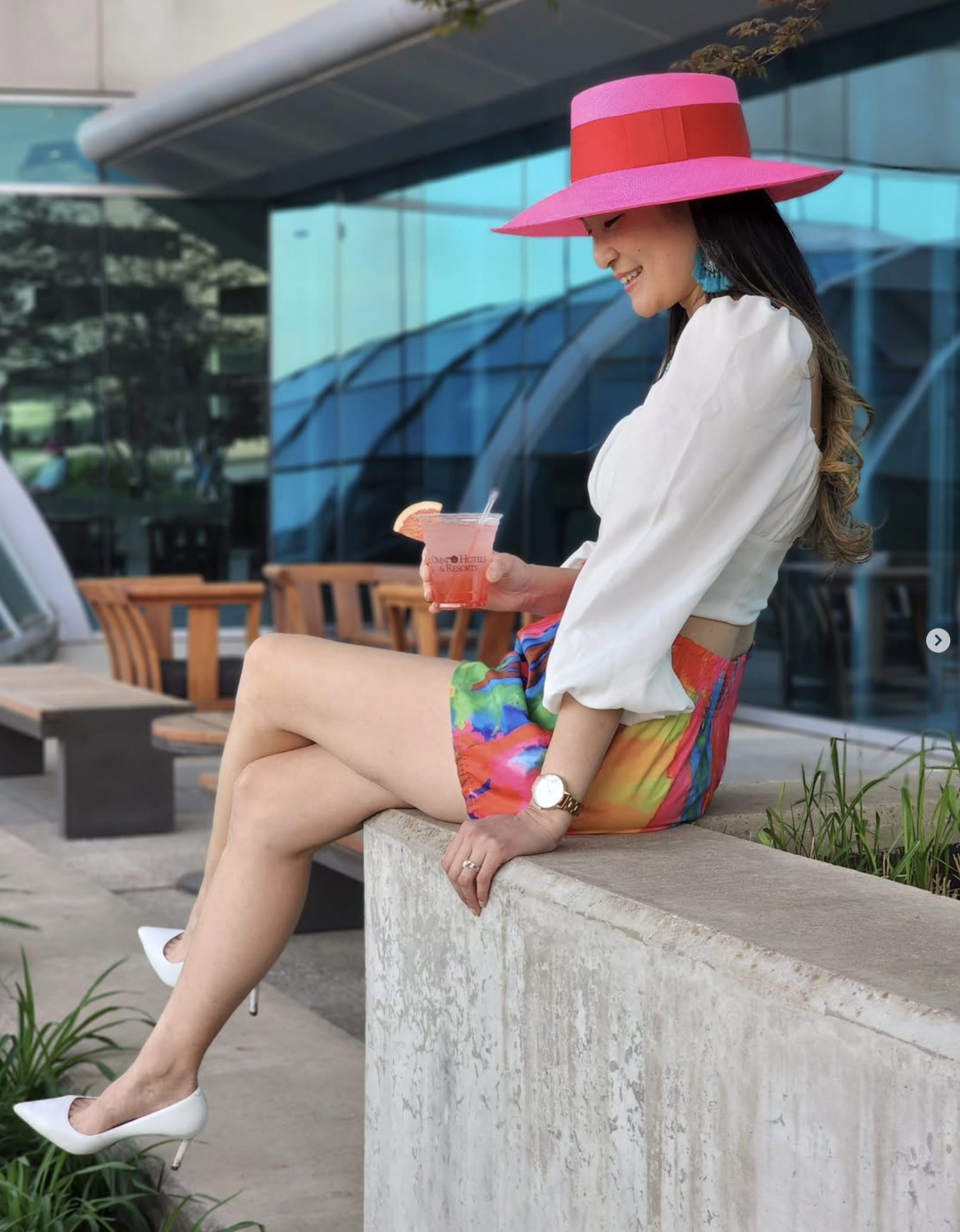 Woman sitting on a concrete ledge outdoors, wearing a pink large-brimmed hat, white blouse, multicolored shorts, white high heels, and holding a pink cocktail with a slice of lemon. She is smiling and looking downward.