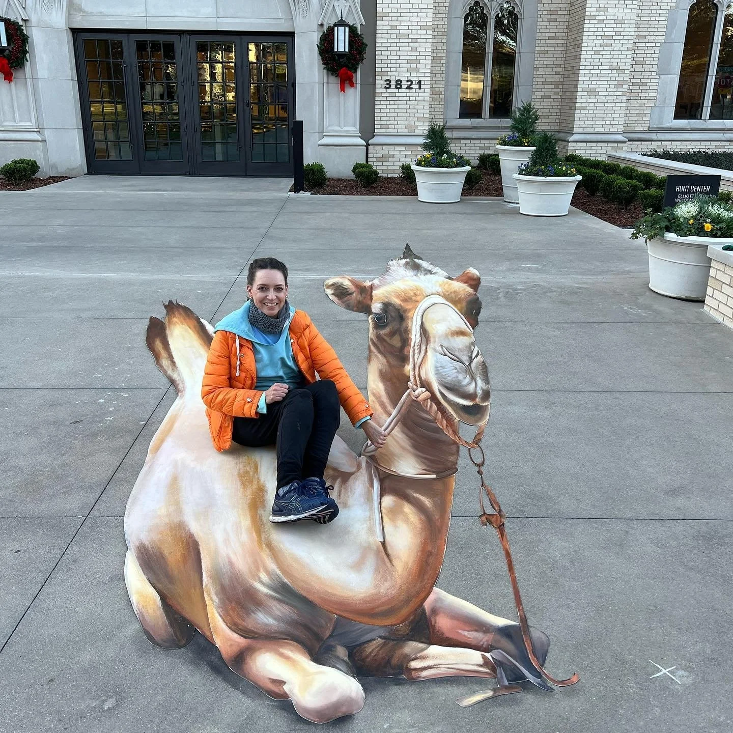 A woman sitting on a painted cutout of a camel with a woman riding it in front of a building with a glass door and holiday wreaths.