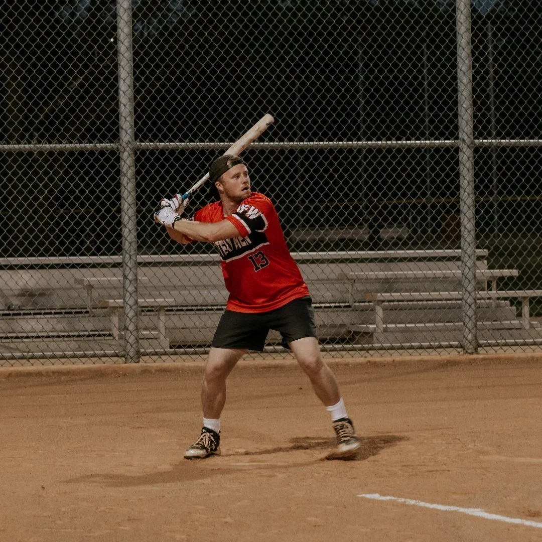 A baseball player in a red jersey and black shorts is preparing to bat on a dirt field at night, with a chain-link fence in the background.