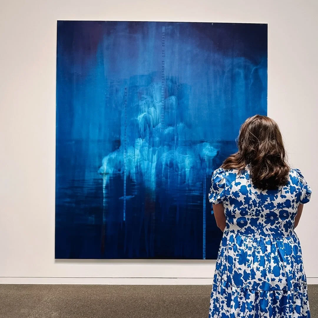 A woman in a blue and white floral dress viewing a large abstract blue painting in an art gallery.
