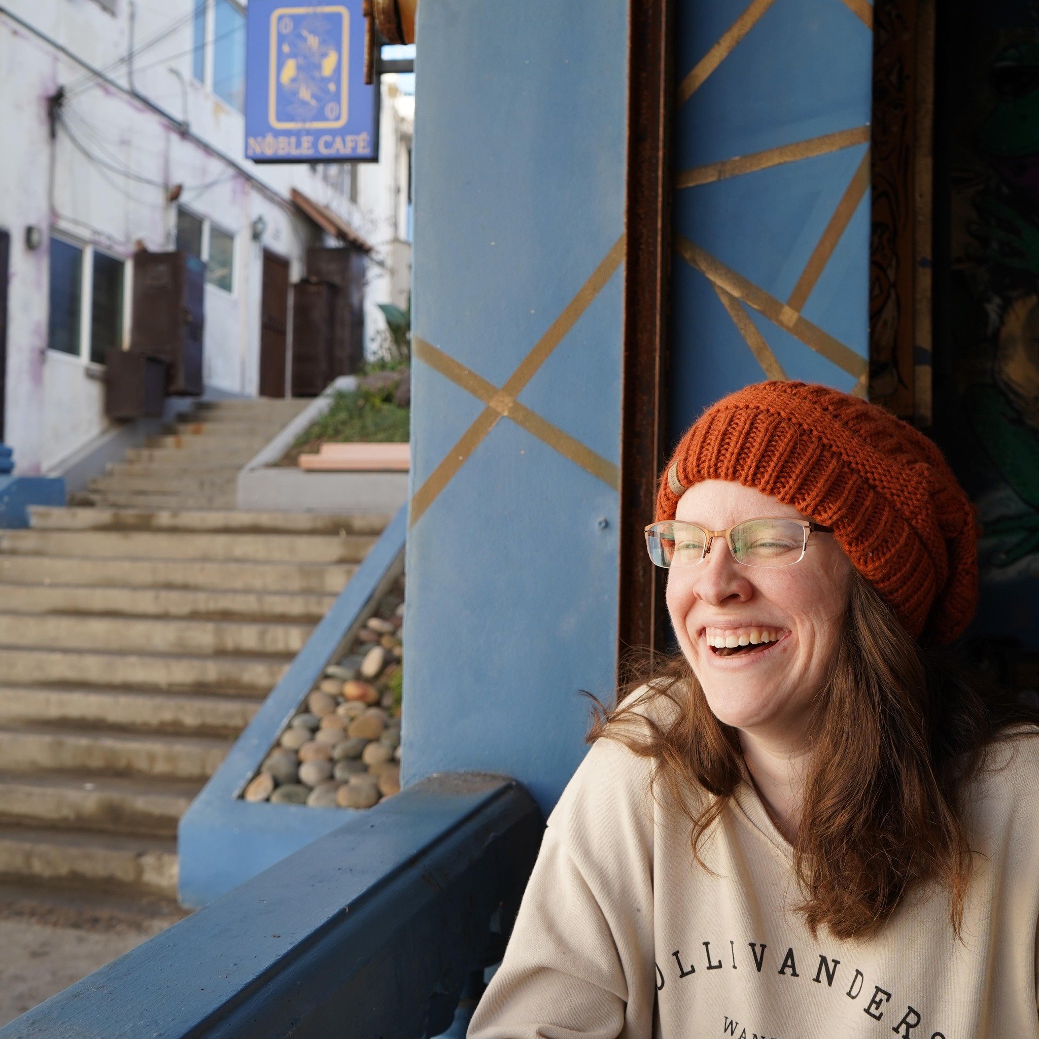 A woman with glasses and a red knit hat laughing happily, sitting near a blue painted wall with gold stripes, with a staircase and a sign for Noble Café in the background.
