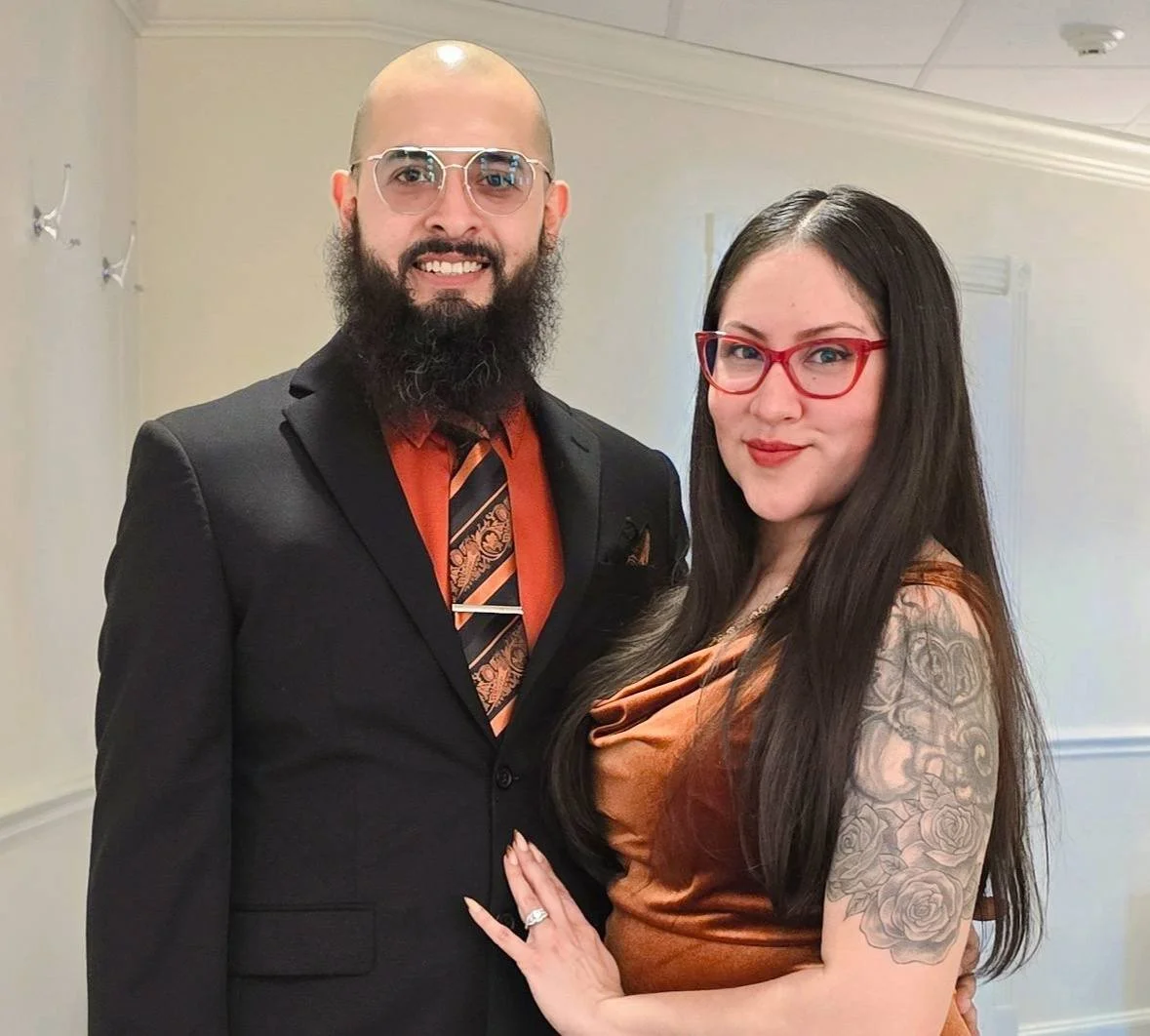 A man with glasses, a beard, and a bald head wearing a black suit and a patterned tie, standing next to a woman with long dark hair, glasses, and a tattoo on her right arm, both smiling and posing indoors.