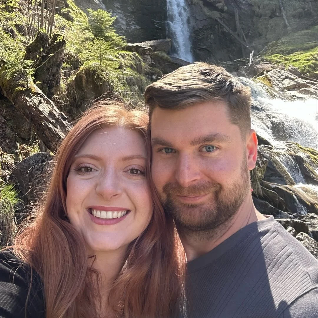 A smiling couple taking a selfie outdoors near a waterfall with rocks and trees in the background.