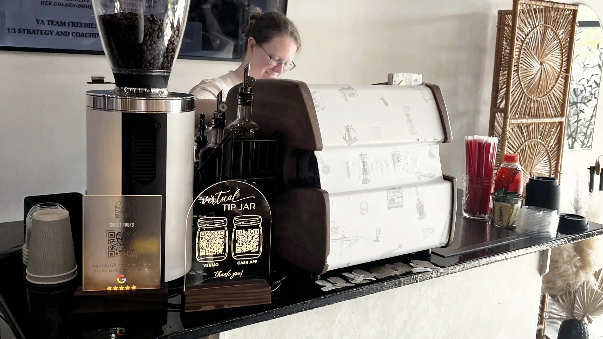 A barista working behind a mobile coffee cart with various coffee supplies, a mahlkonig espresso grinder, and la spaziale lucca mini a53 espresso machine, in a venue decorated with wicker room dividers and artwork.
