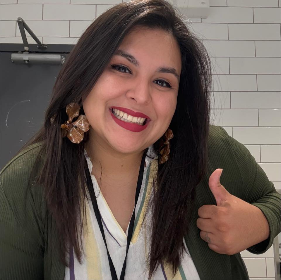 A woman with long dark hair wearing a green jacket and large brown earrings, smiling and giving a thumbs-up gesture, standing in front of a tiled wall.