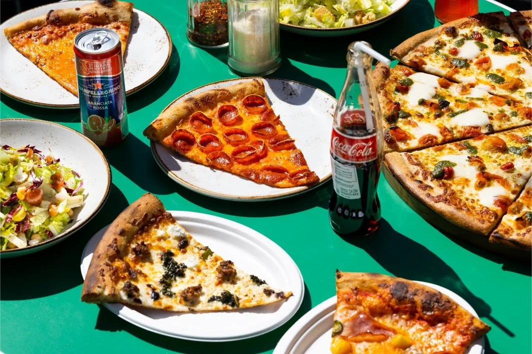 Several slices of pizza on white and patterned plates, a salad in a bowl, a can of sparkling water, and a bottle of Coca-Cola on a green table outdoors.
