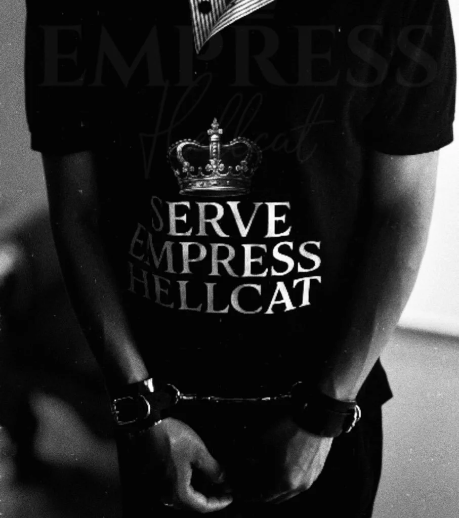 Person wearing a black T-shirt with a crown graphic and the phrase "Serve, Empress, Hellcat" in bold white letters, handcuffed with hands in front of them.