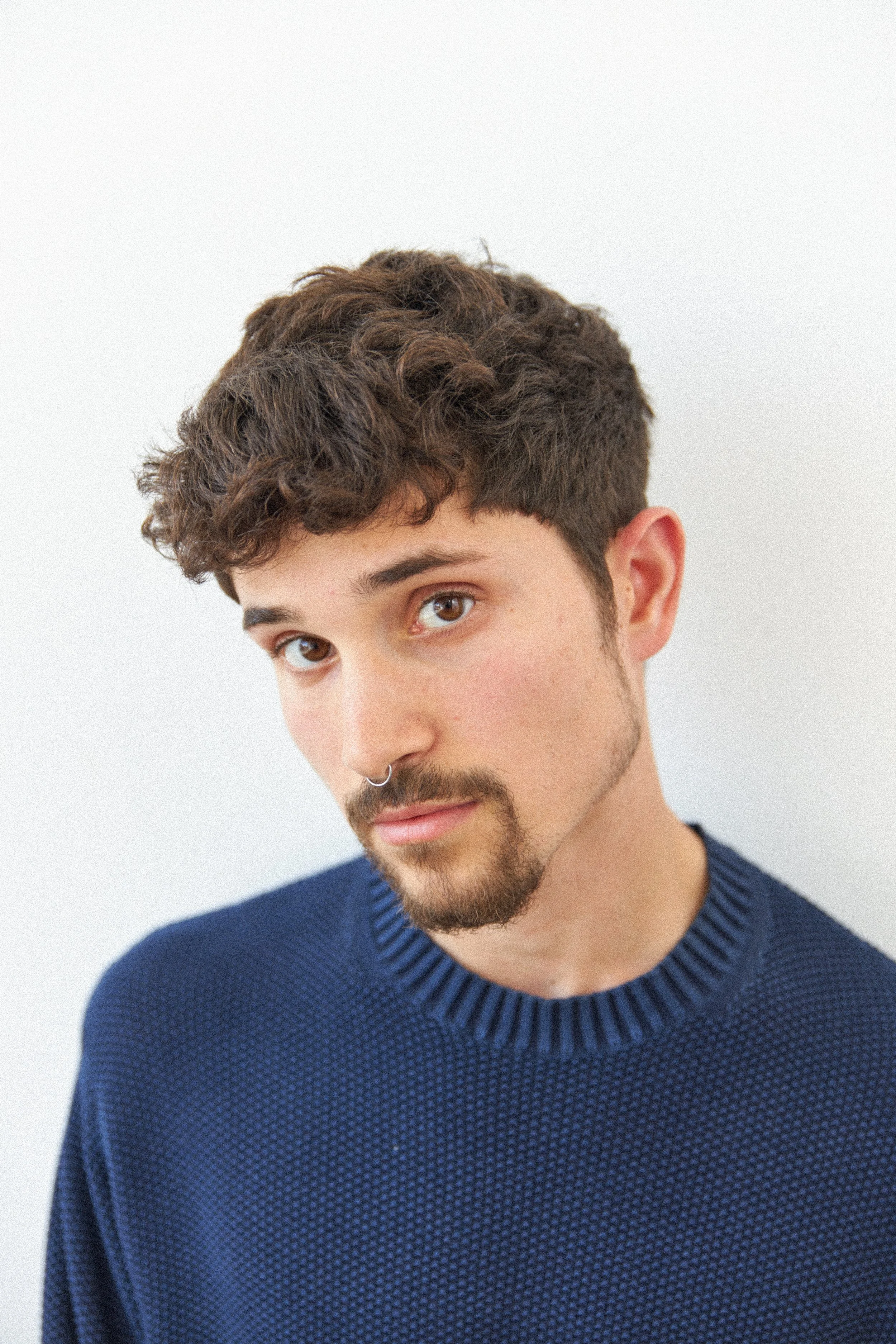 A young man with brown eyes, curly brown hair, and a nose ring, wearing a blue textured sweater, looking at the camera.