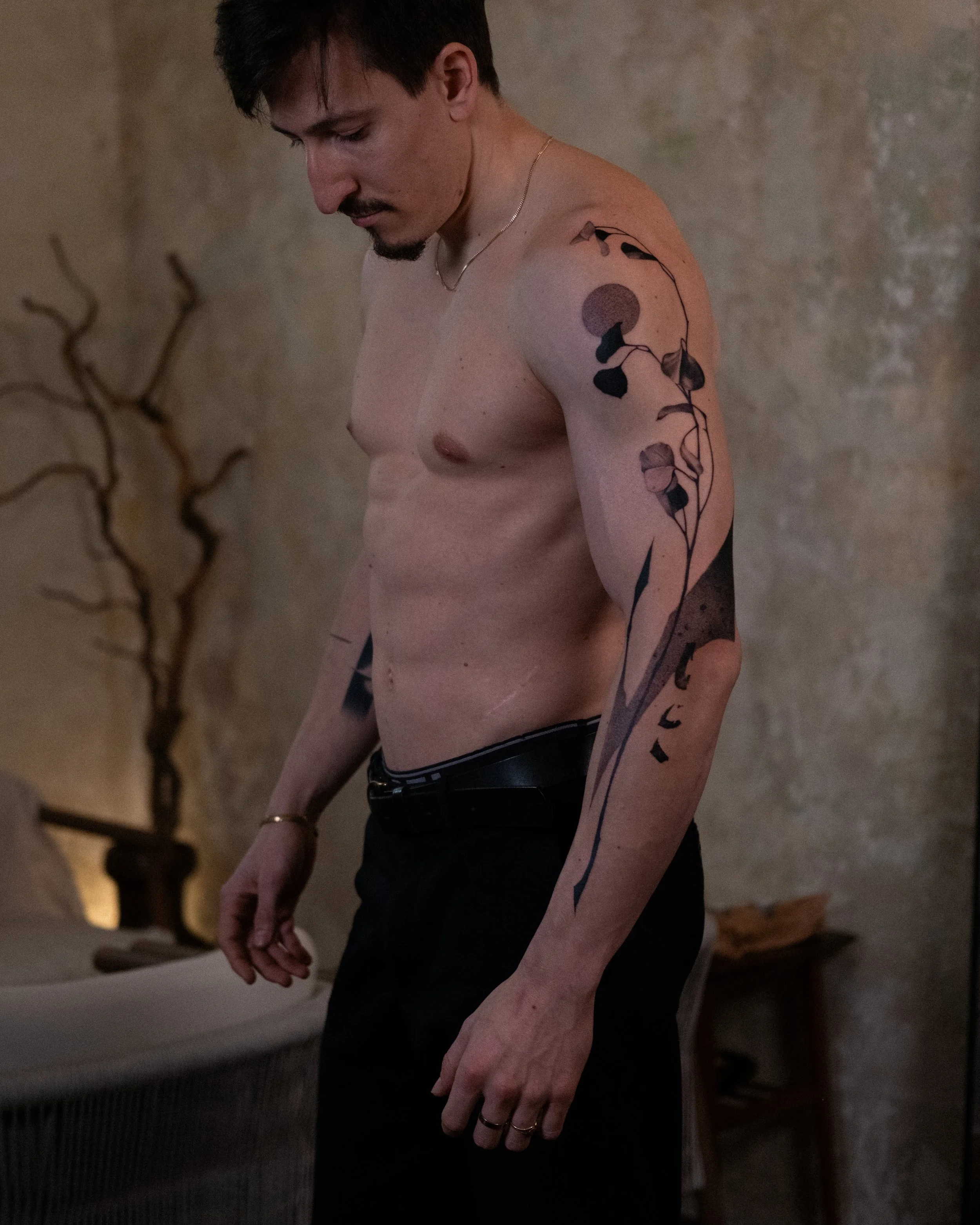 A shirtless man with tattoos on his arm, looking downward, standing in front of a beige wall with a textured surface. The tattoos include a black and grey plant design and a circular shape.