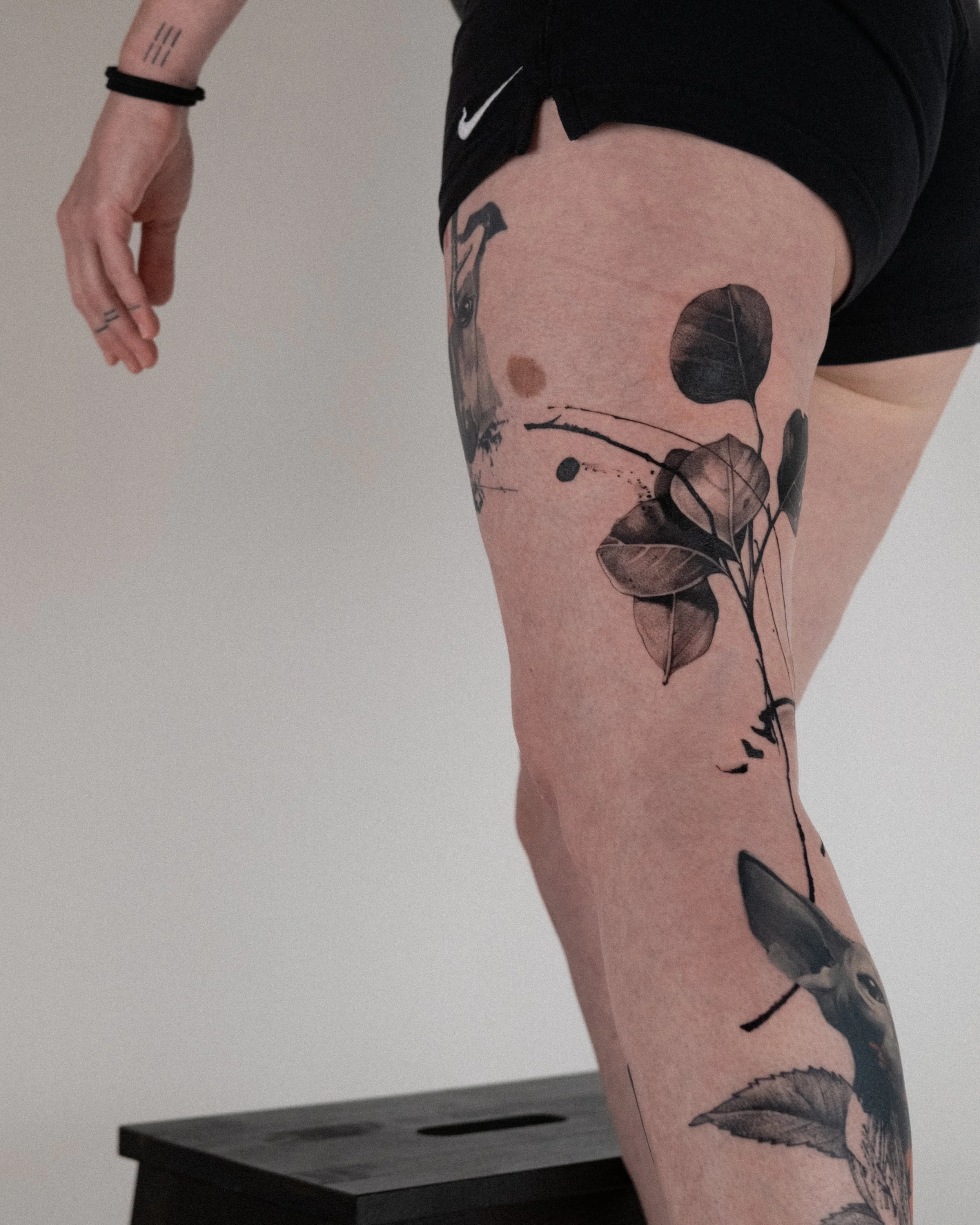 Close-up of a person's upper arm and thigh, showing black tattoos of leaves and abstract designs, wearing black shorts with a Nike logo.
